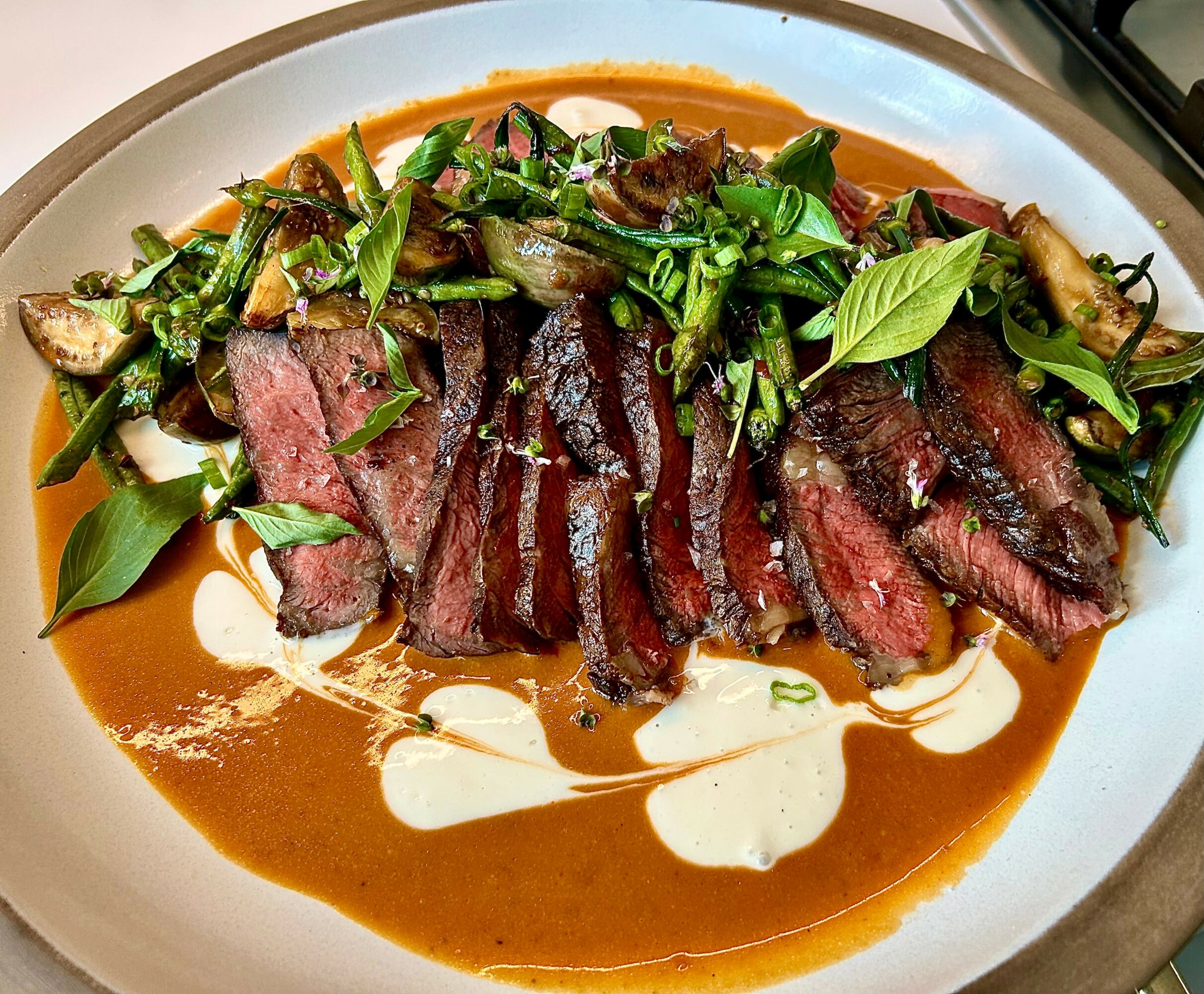 Sliced cooked steak with green vegetables, herbs, and sauces on a white plate.
