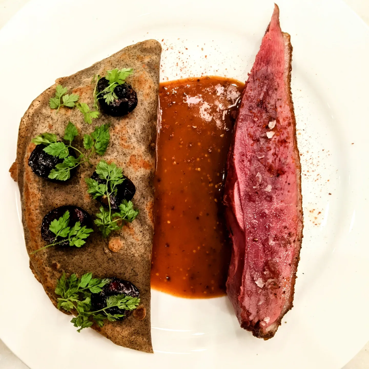 Plate with a slice of cooked meat, sauce, and a folded flatbread garnished with herbs and black olives.