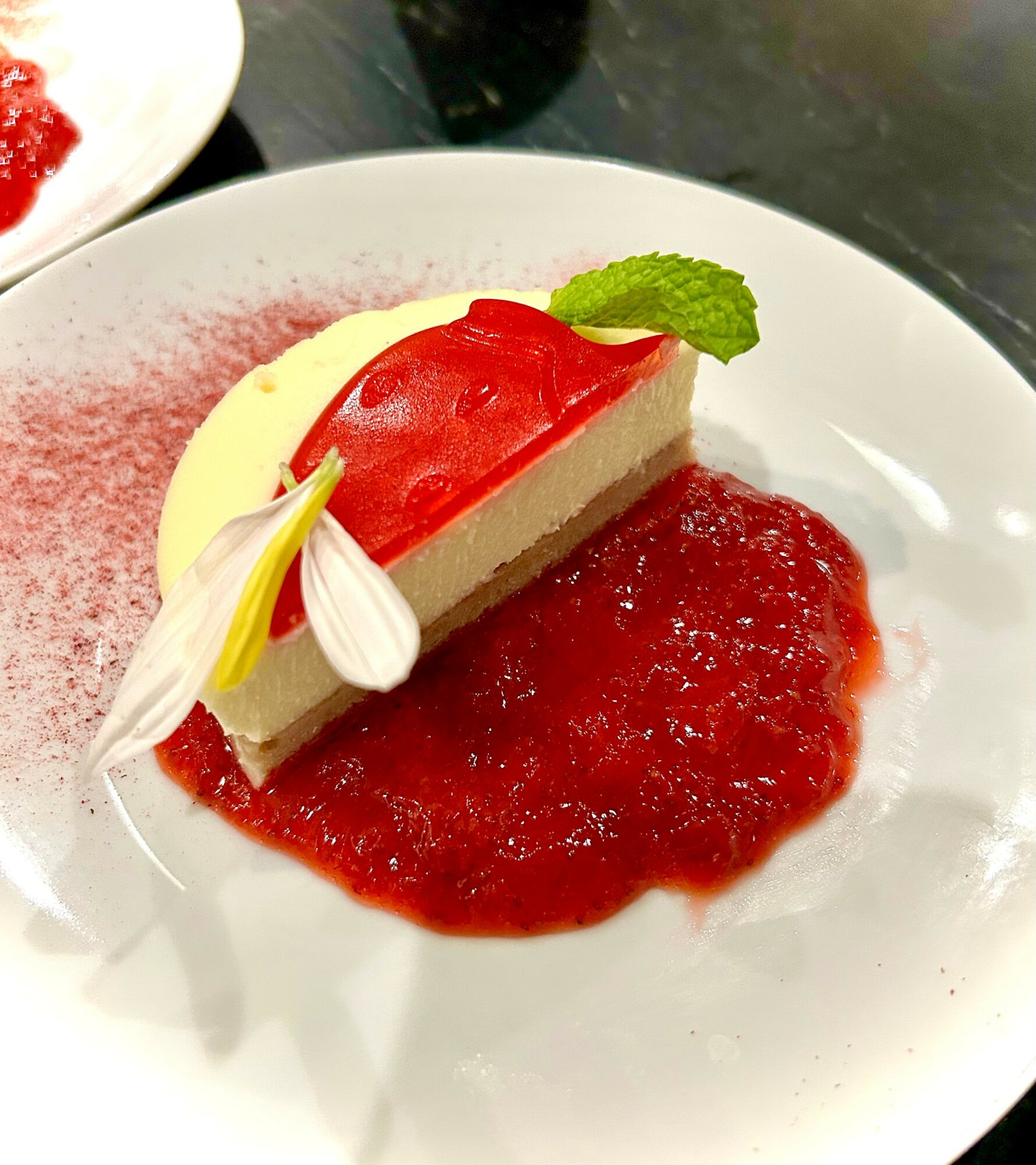 Cheesecake with red fruit topping, garnished with a mint leaf and edible flower, on a white plate with red sauce, 20 words