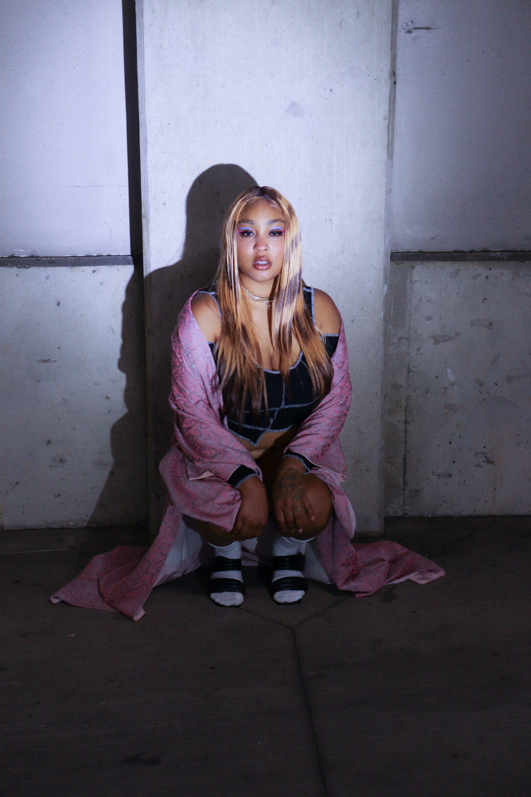 Young woman with long hair sitting against a concrete wall, wearing a pink jacket, striped socks, and black dress.