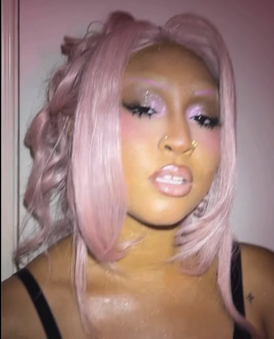 Woman with pink hair, makeup, and glittery eye makeup, looking at camera with slightly open mouth.