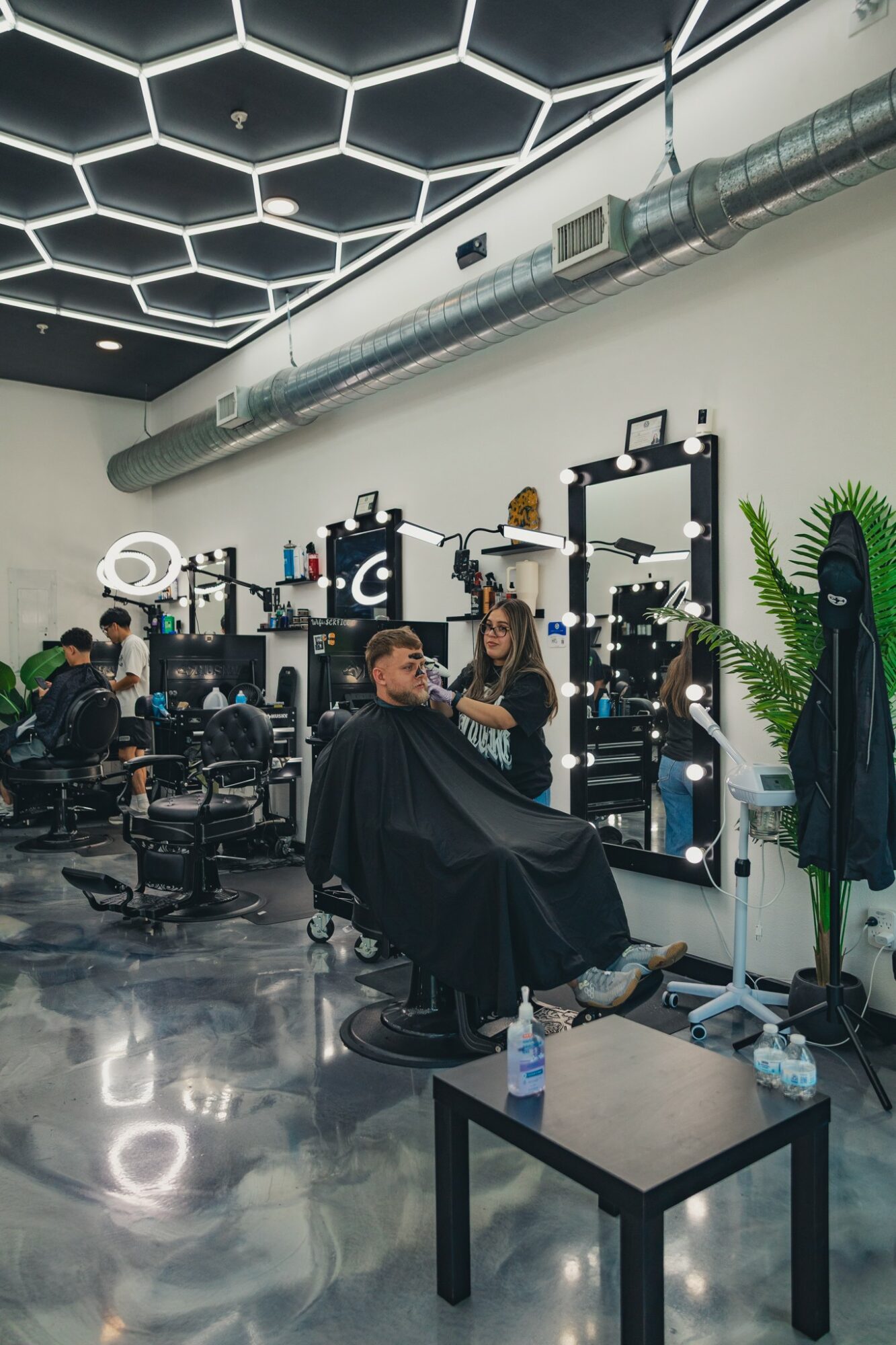 Person getting a haircut in a modern salon with bright lighting and multiple styling stations, people working and waiting.