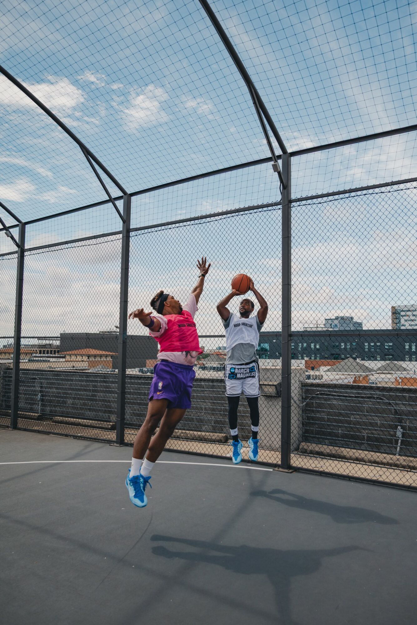 Two children playing basketball on an outdoor court with a chain-link fence, sky and buildings in background.