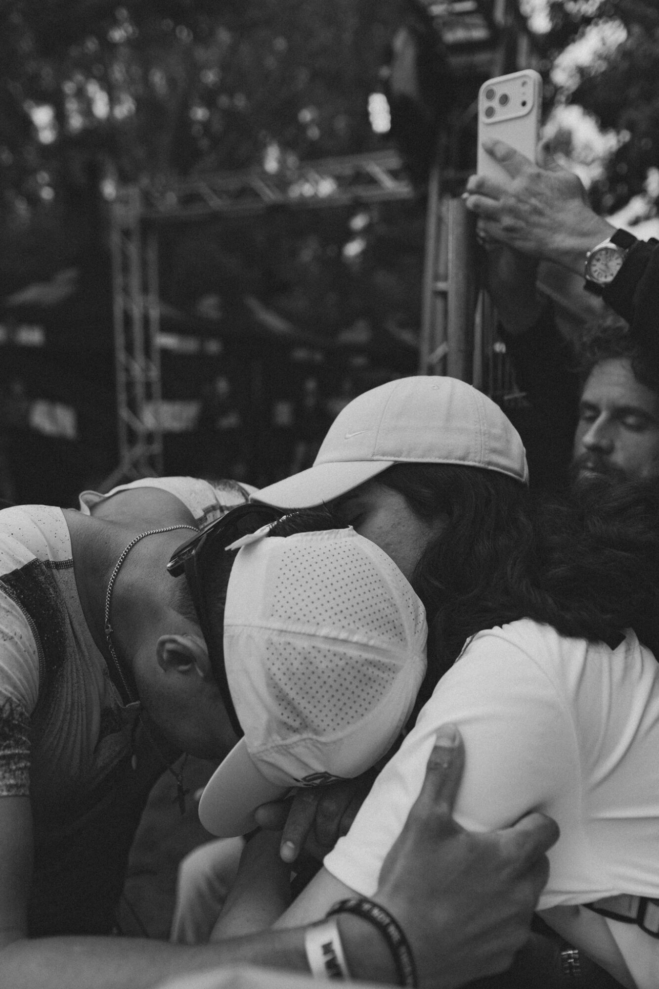Group of people hugging outdoors, one person wearing a white cap, others embracing, person taking photo in background.
