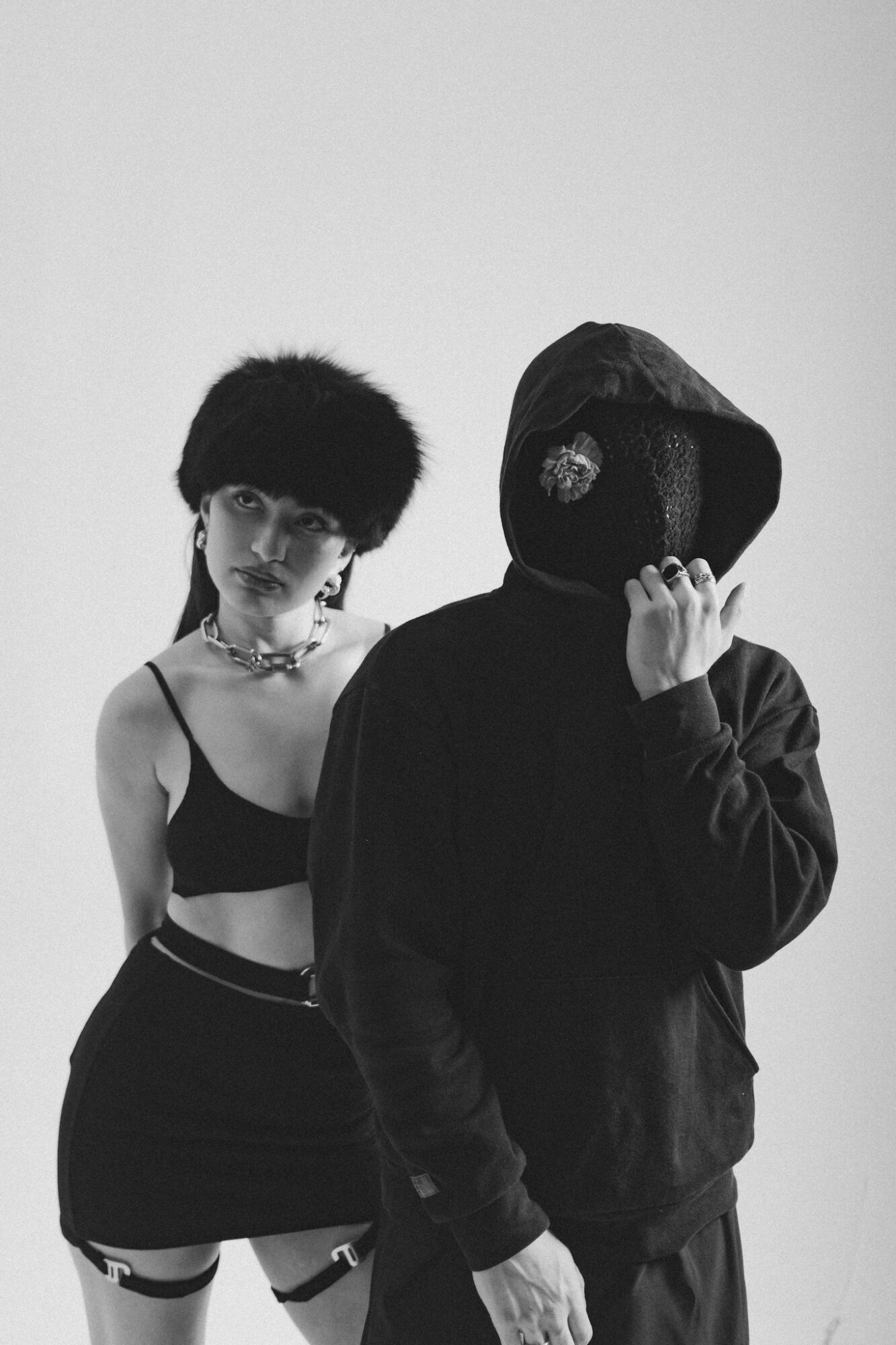 Two people pose against a plain background, one with a hood covering their face, the other with short curly hair and a crop top.