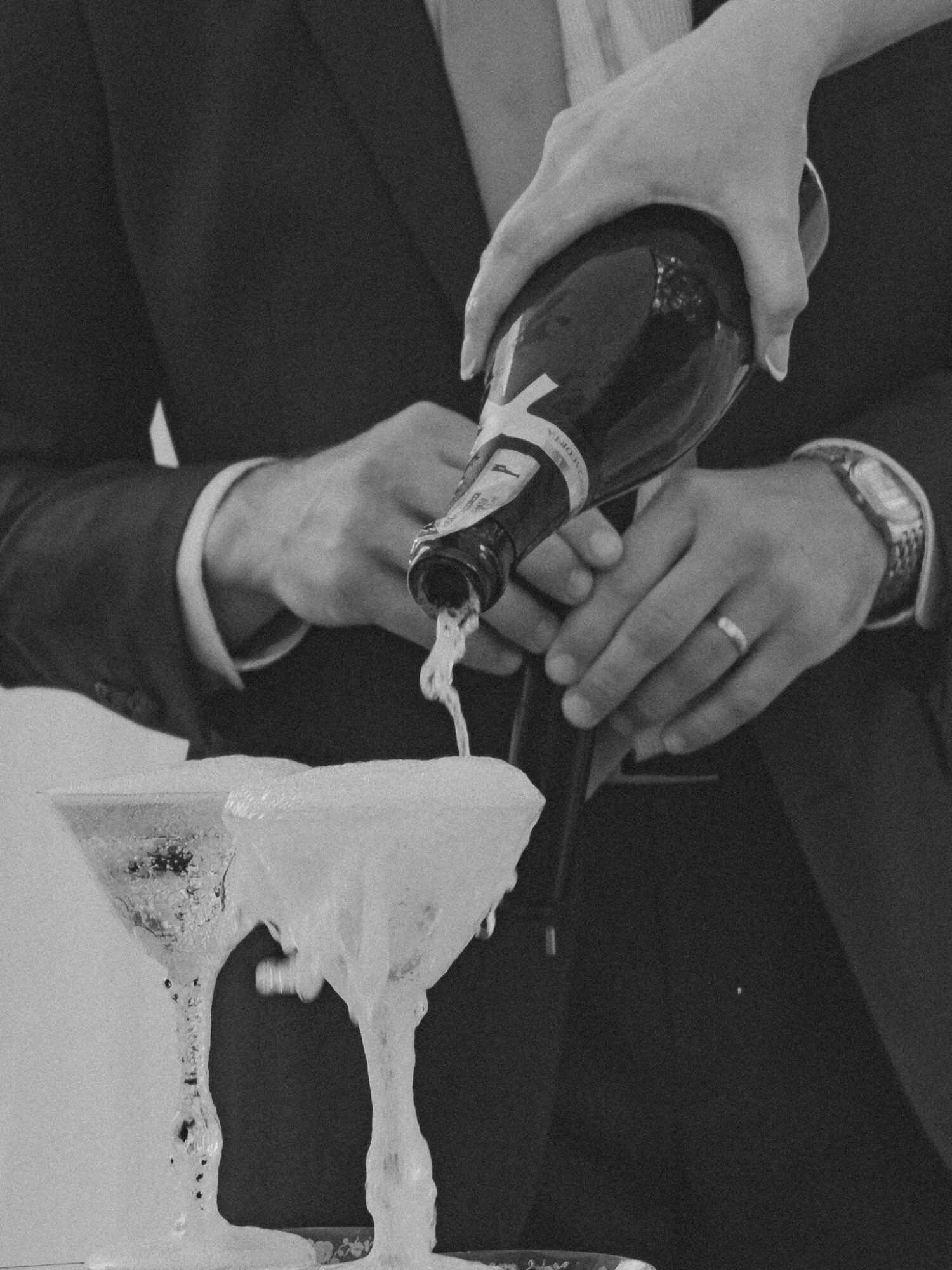 Two people pour champagne into a glass, with one holding the bottle and the other supporting the glass, in black and white.