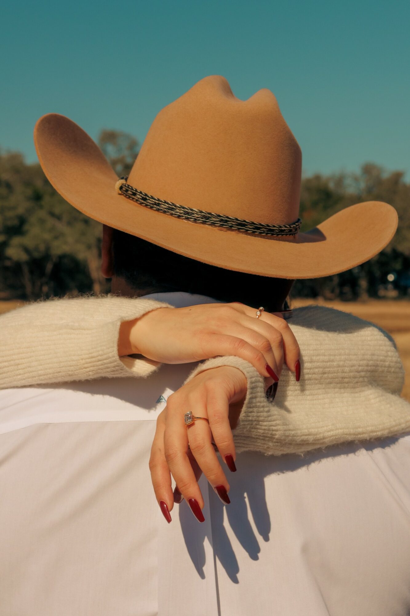 Person wearing a wide-brimmed hat and cream sweater, resting their arms on a surface outdoors with trees in background.