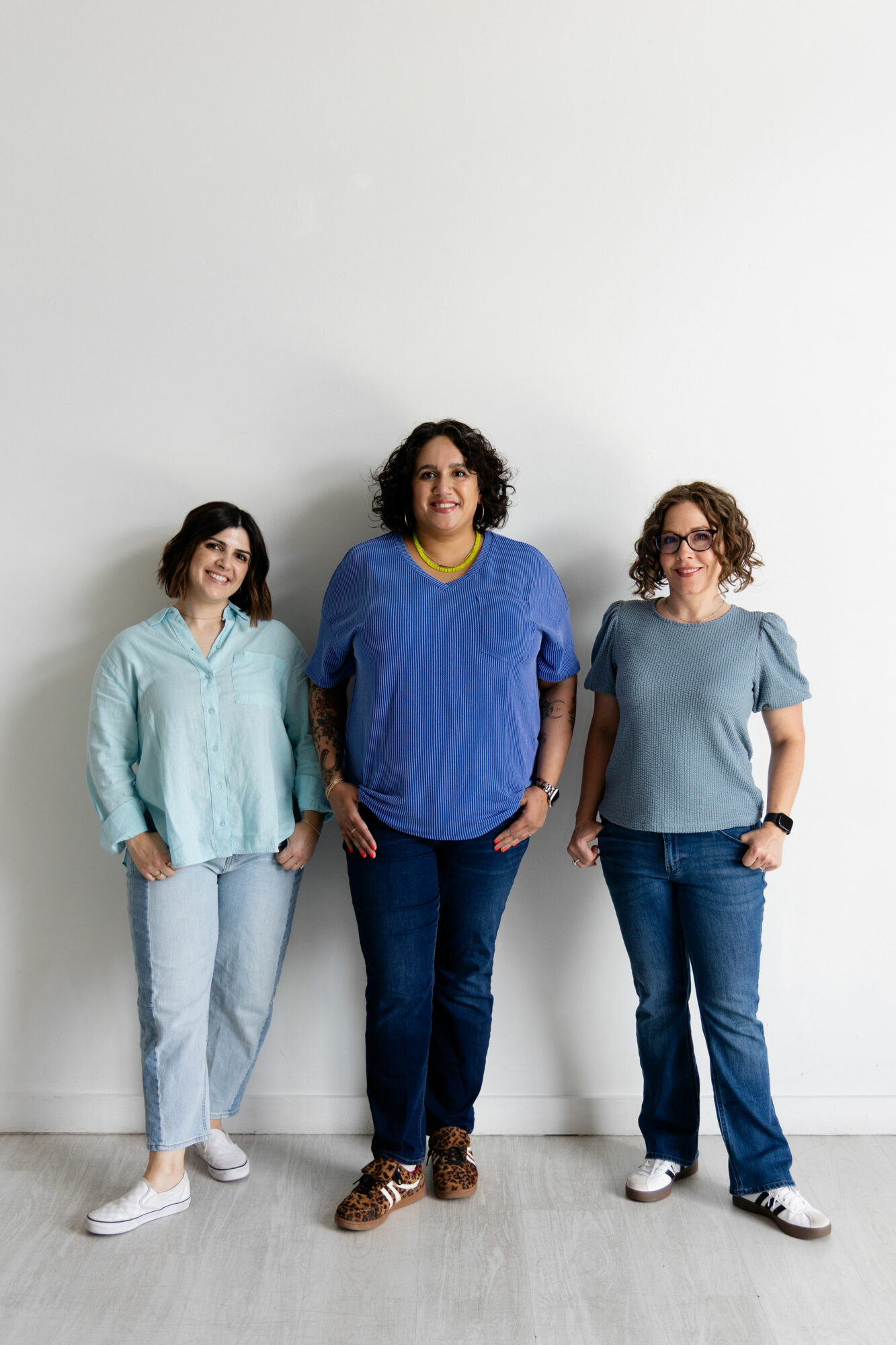 Three women standing against a plain white wall, smiling, wearing casual clothing, with a light-colored floor.
