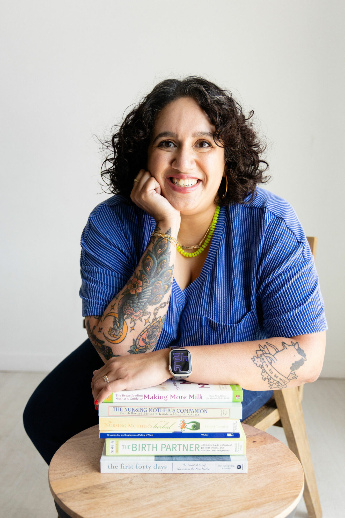 Woman with curly hair smiling, resting her chin on her hand, wearing a blue shirt, sitting at a table with books.