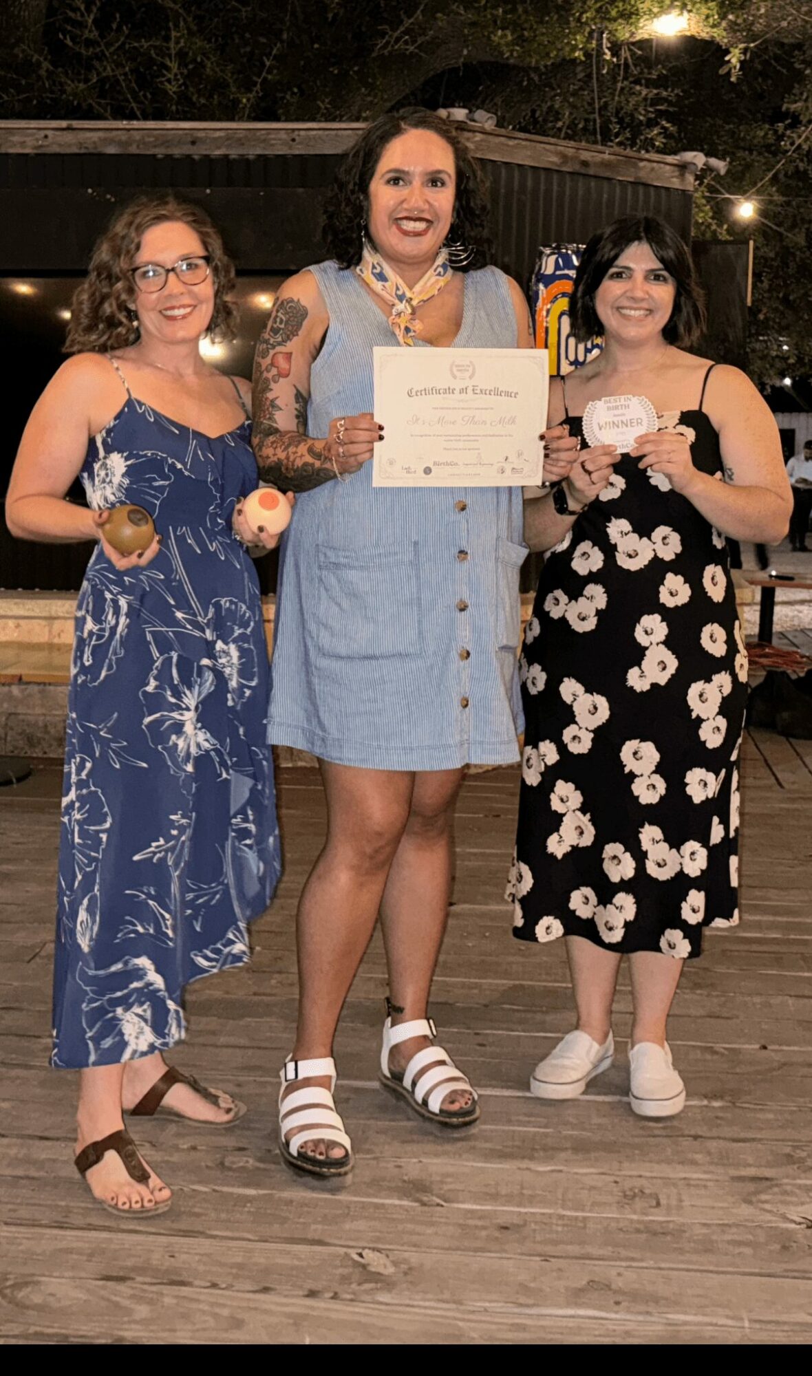 Three women standing outdoors at night, one holding a certificate, all smiling, dressed in summer clothing, on a wooden surface.