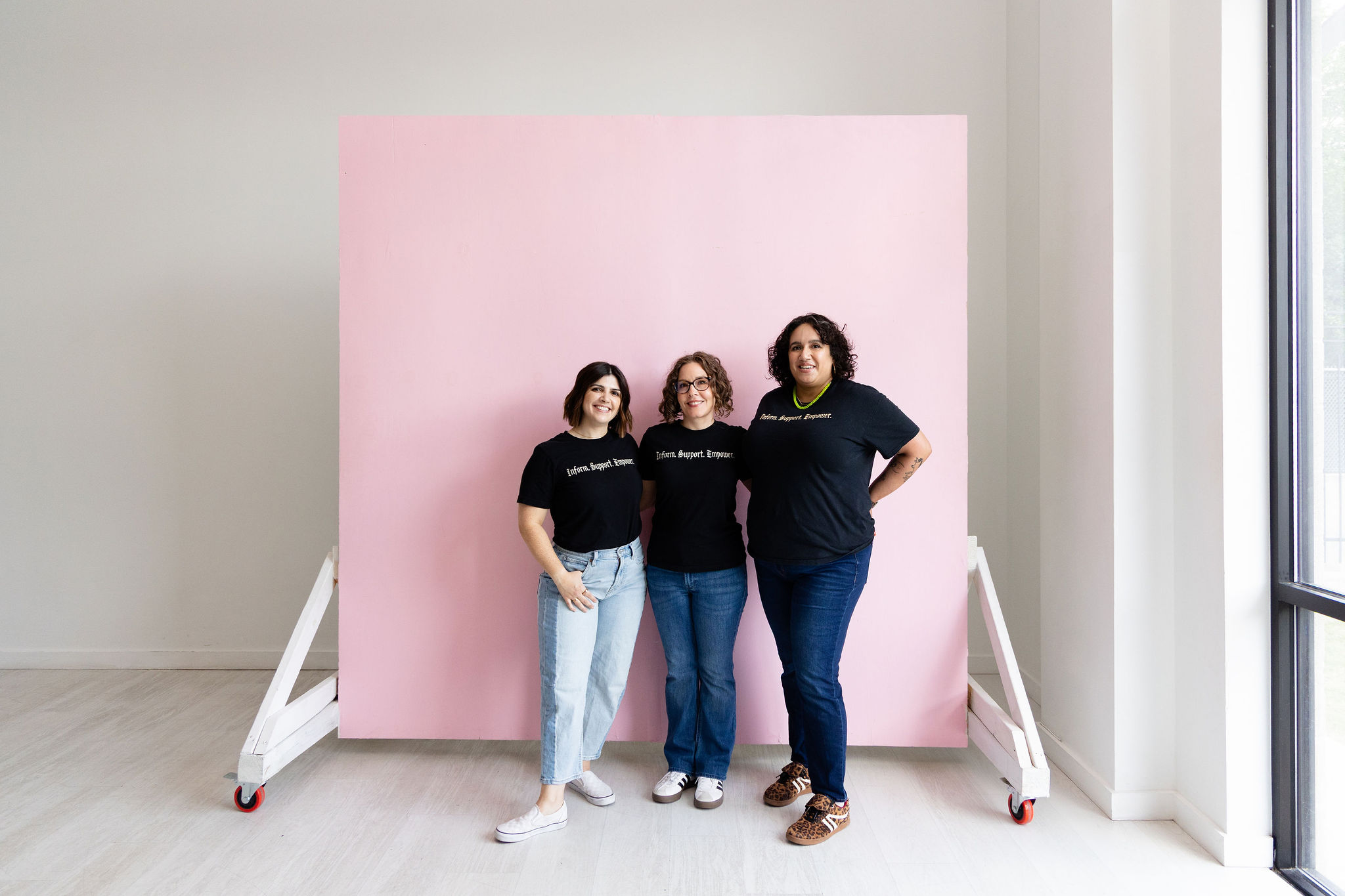 Three women stand in front of a pink backdrop in a bright room with large windows, smiling at the camera.