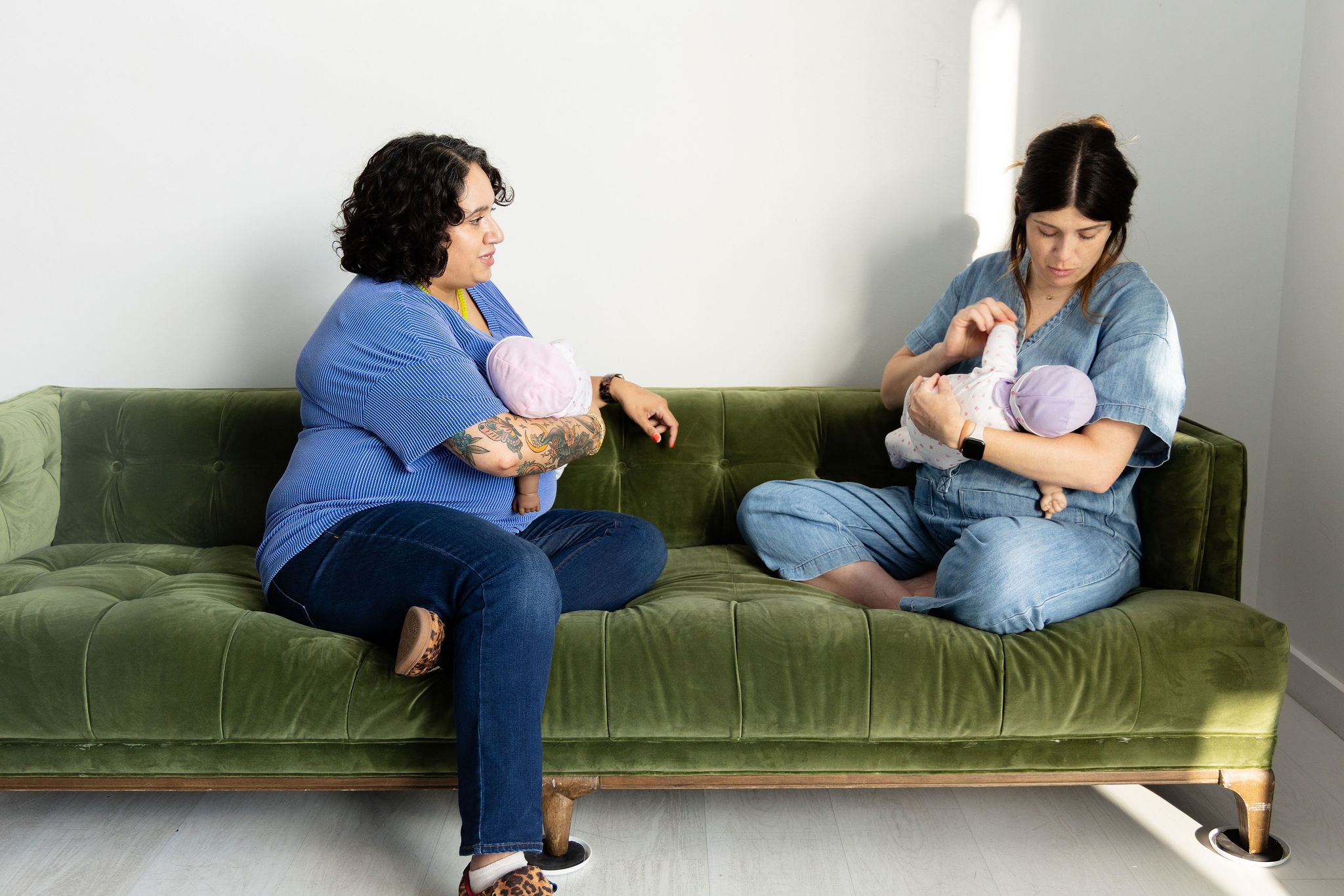 Two women sitting on a green sofa, each holding a baby wrapped in a blanket, in a bright room.