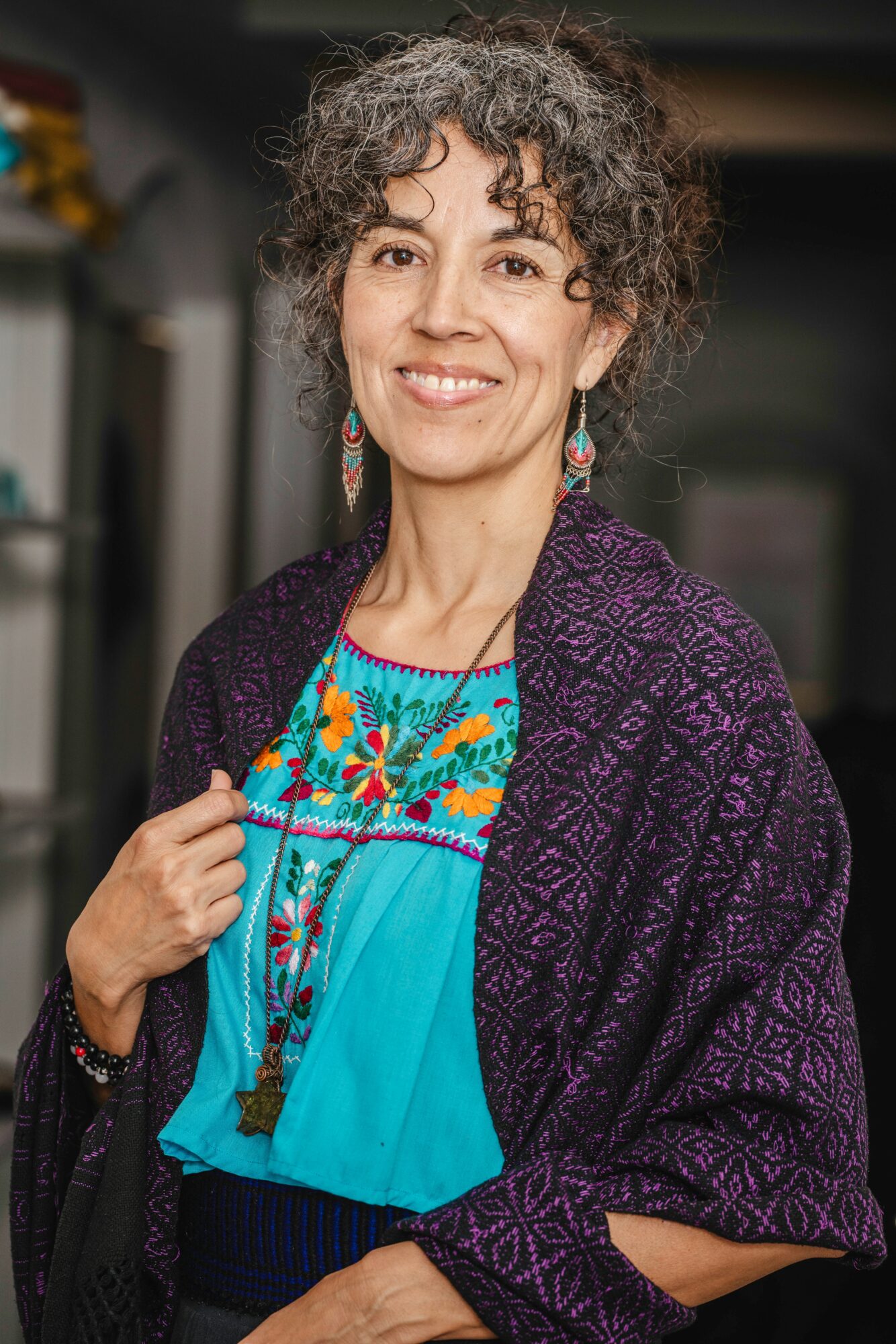 Woman with curly hair smiling, wearing colorful embroidered blouse and purple patterned shawl, standing indoors.
