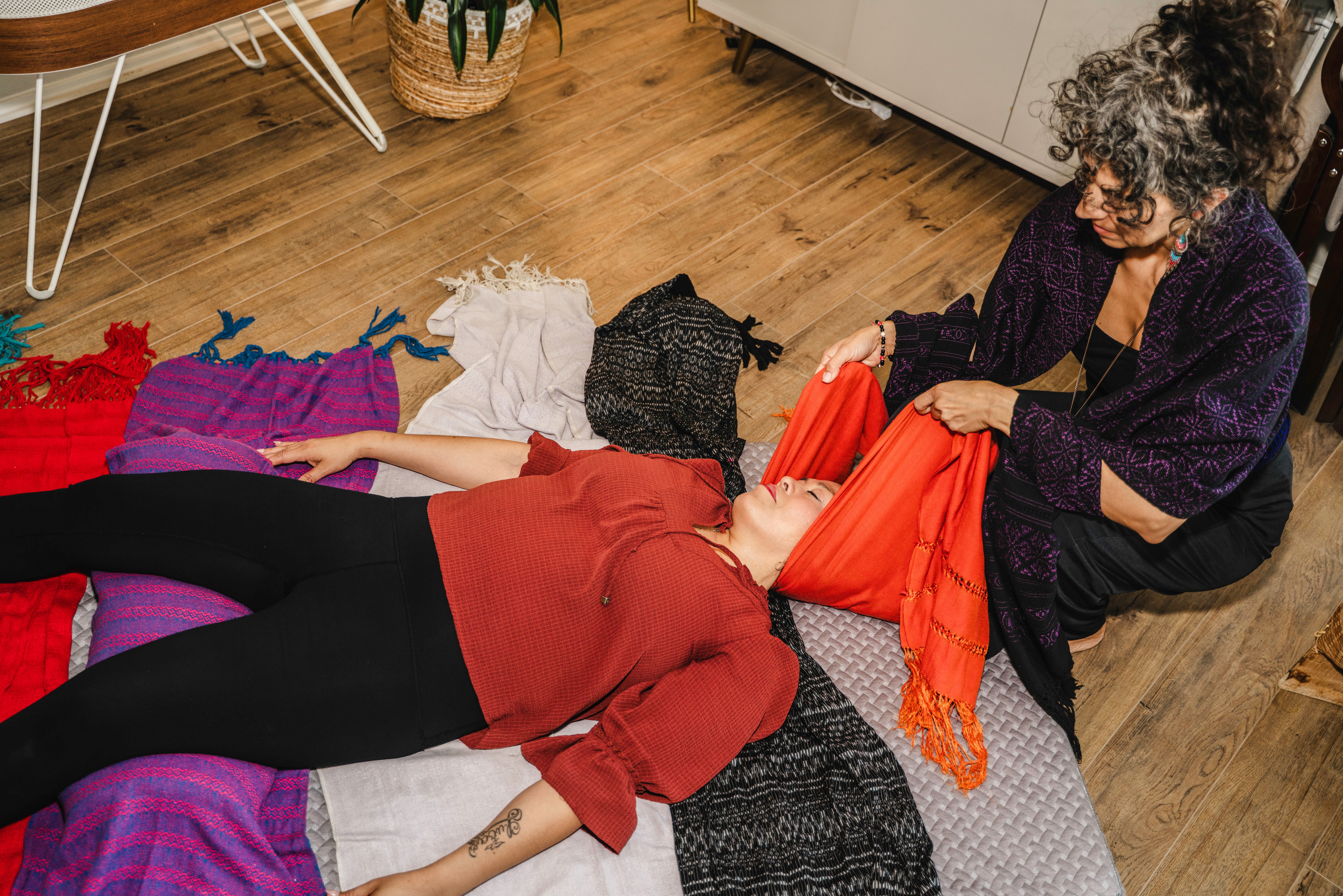 Two women on the floor with colorful fabrics, one lying down and the other sitting, in a room with wooden flooring.