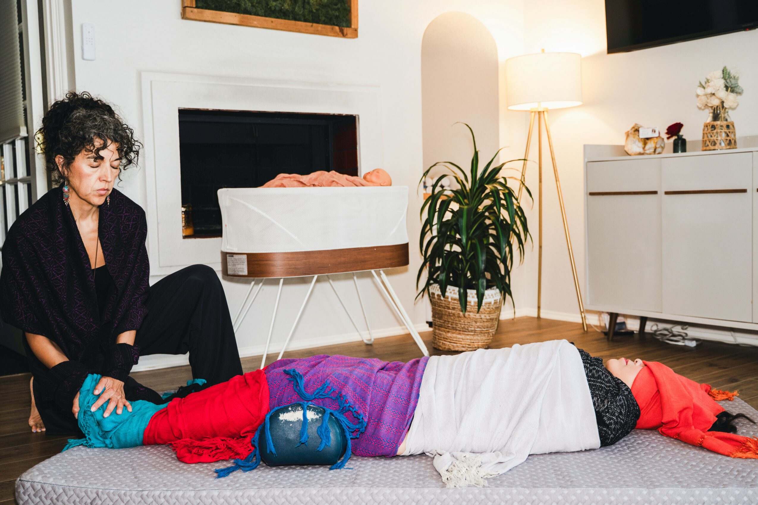 Person lying on the floor with colorful clothing and headphones, another person sitting nearby, living room setting with furniture and plants.