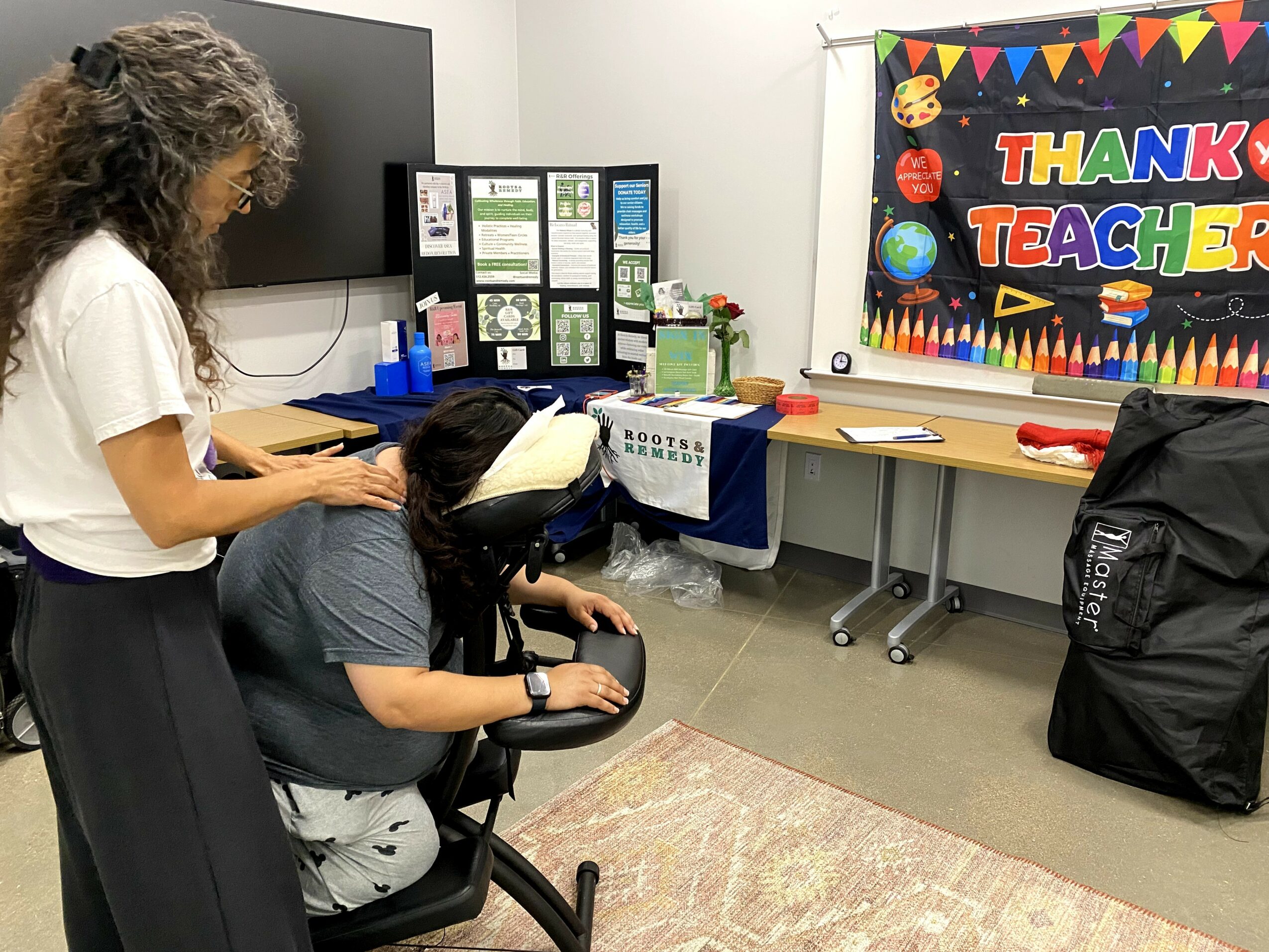 Two women in a classroom, one standing and one sitting, with a colorful 'Thank You Teachers' banner on the wall.