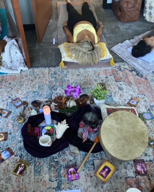 People lying on mats and a person lying on a table, with a decorated table and various items on a patterned rug.