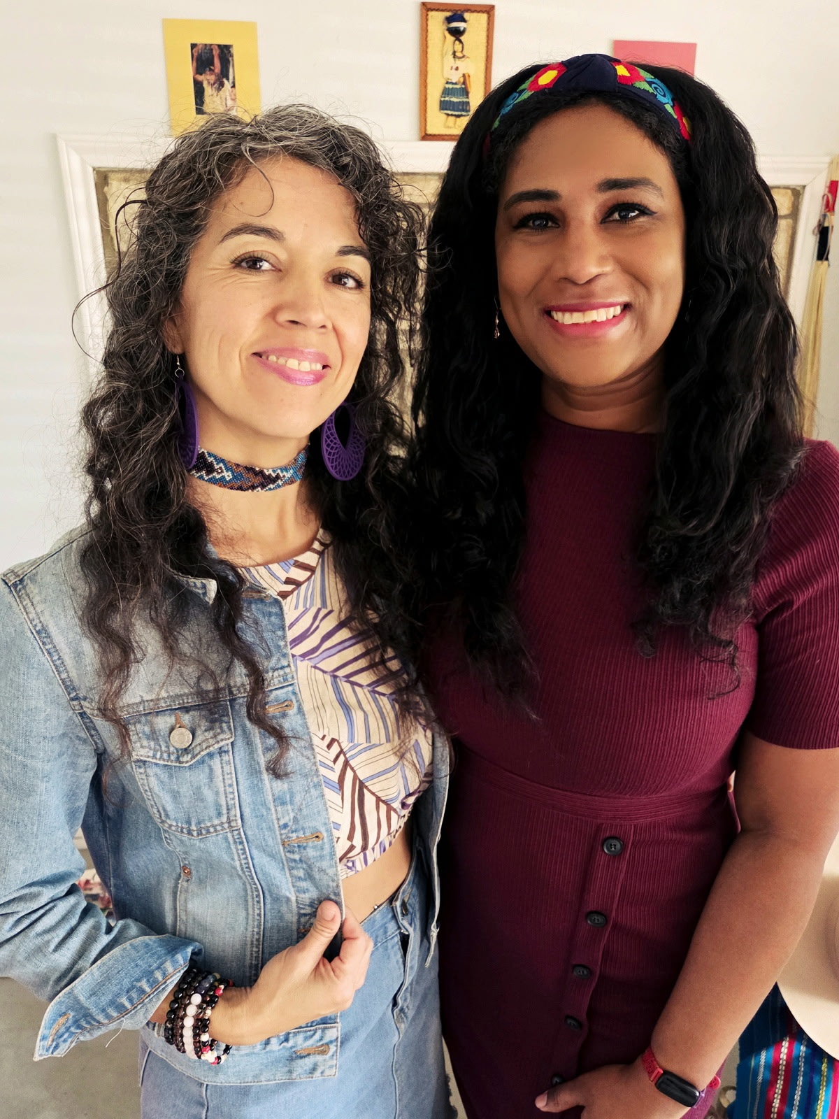 Two women smiling, standing close together indoors with framed pictures on the wall behind them.