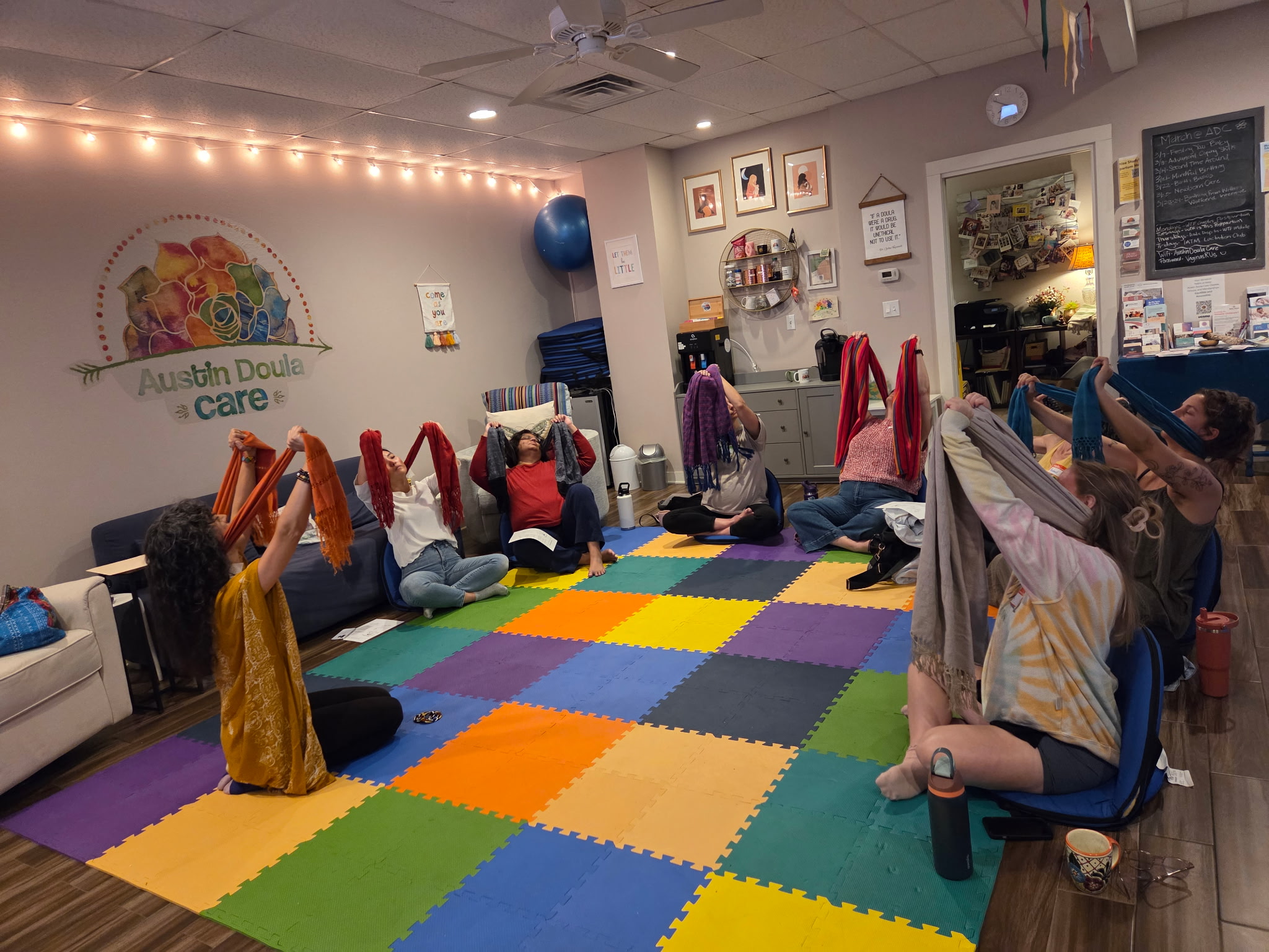 People sitting on chairs in a room with colorful foam mats, holding towels overhead, with a wall decorated with a rainbow and string lights.