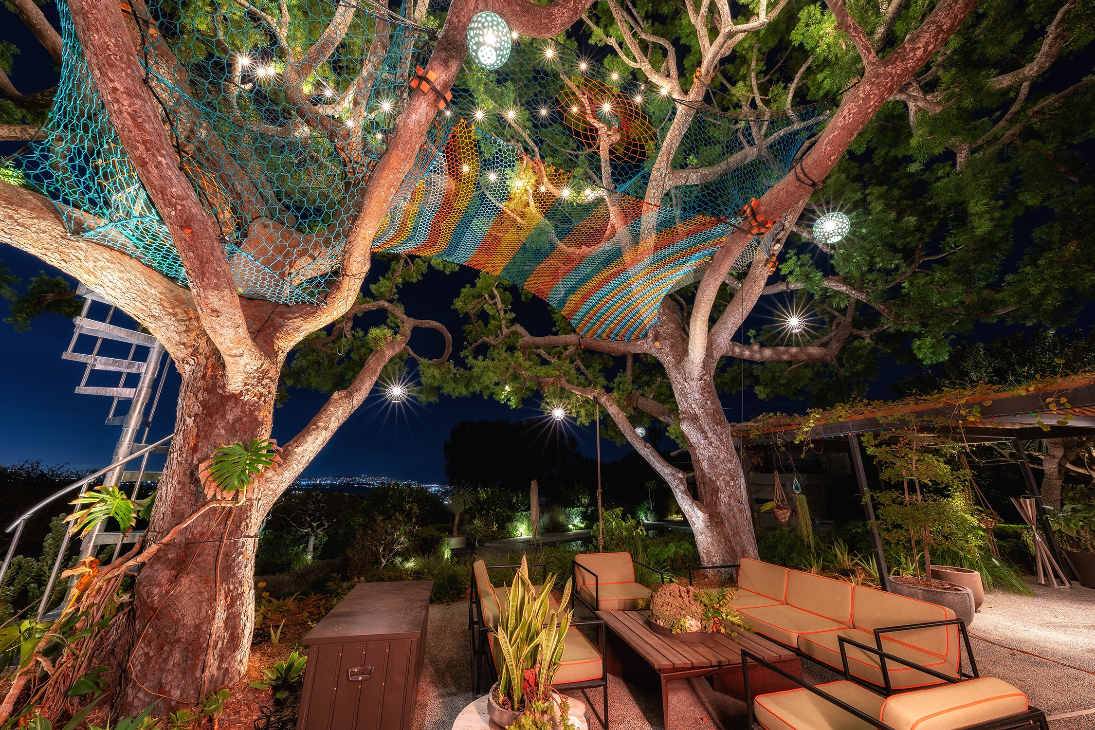 Outdoor seating area under trees with colorful fabric canopy and string lights at night.