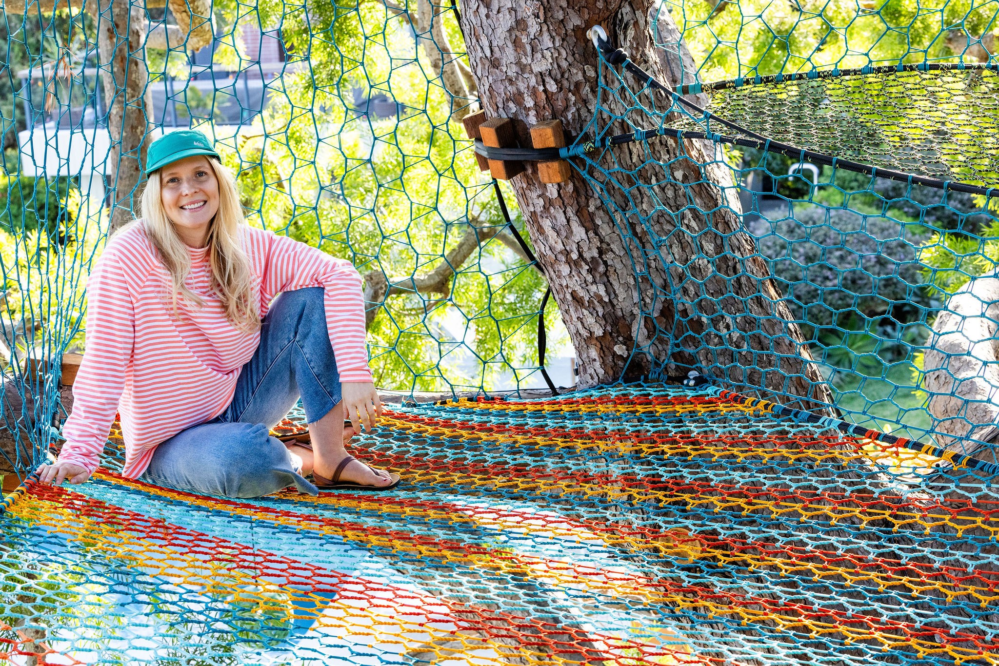 Woman sitting on a colorful hammock outdoors, smiling, wearing a pink striped sweater and a blue cap, surrounded by trees.