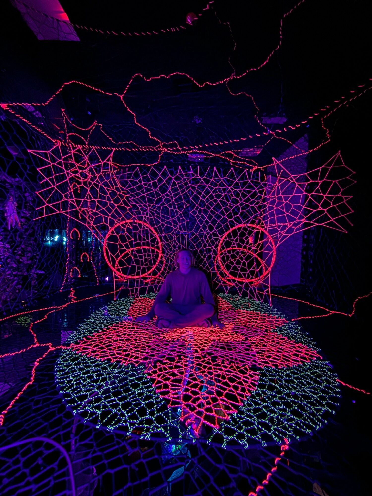 Person sitting on a colorful, illuminated web-like structure with neon lights and abstract patterns in the background.