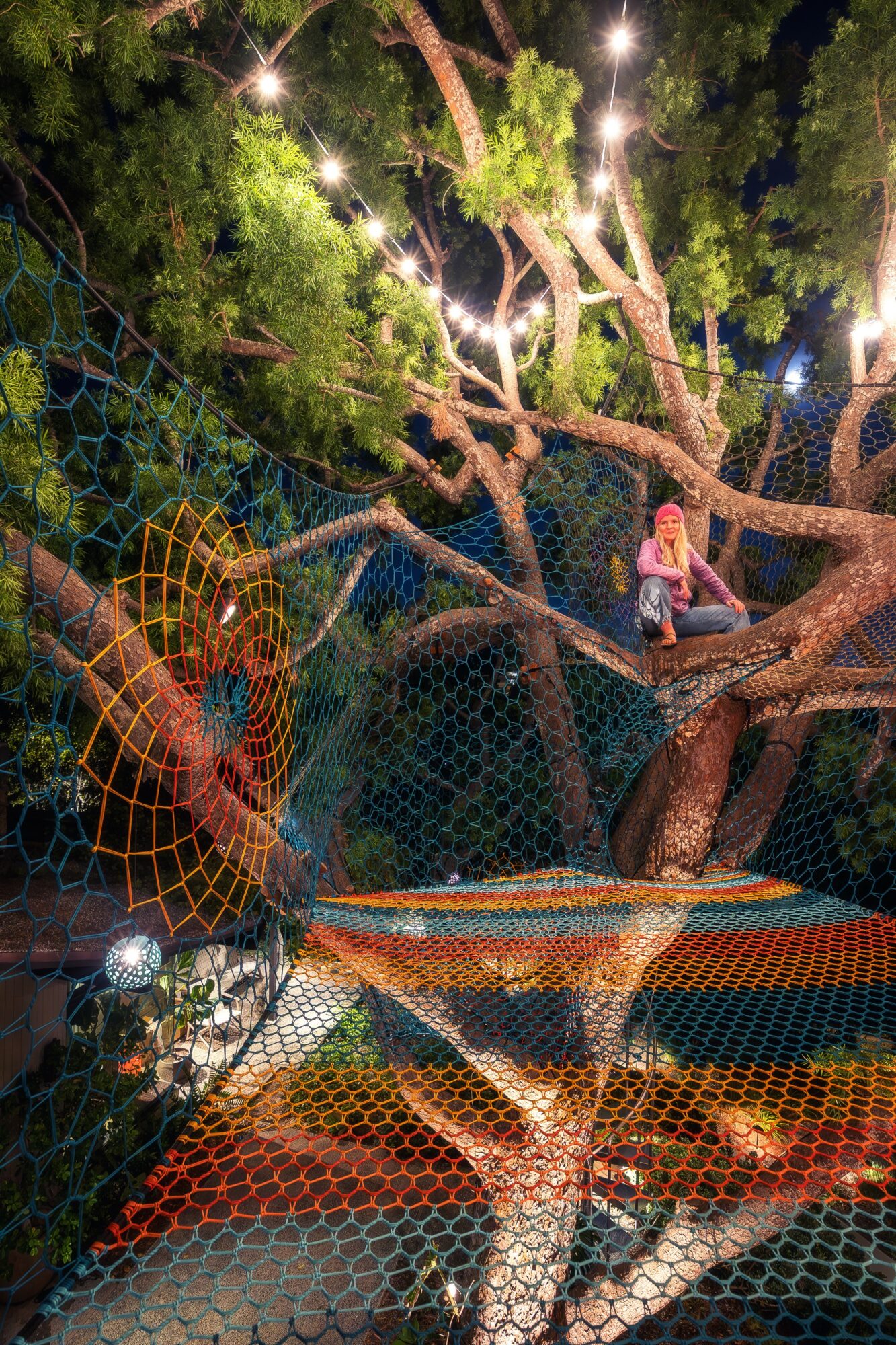 Children playing on tree branches with string lights, surrounded by greenery and a netted area, at night.