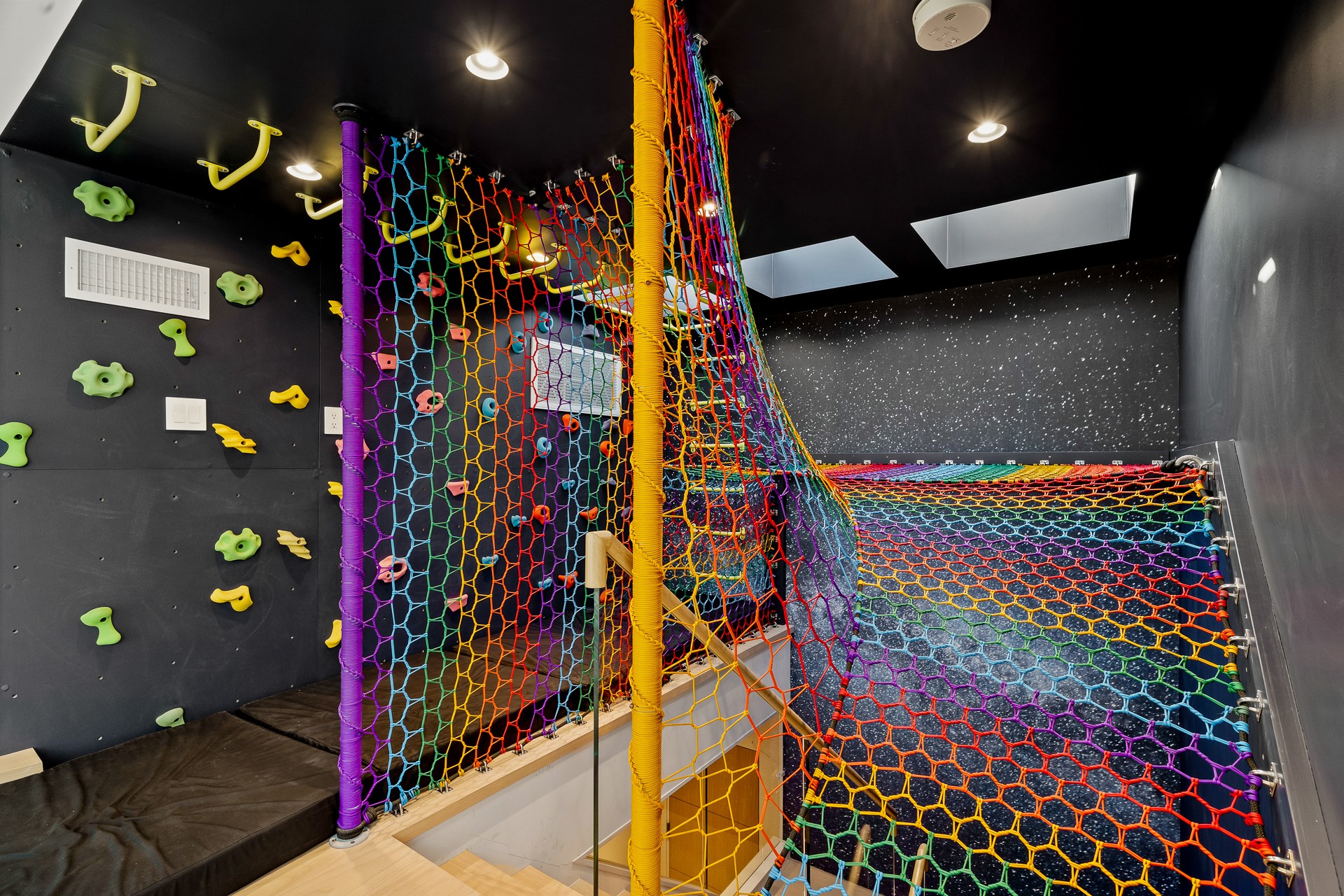 Indoor play area with colorful netting, climbing structures, and a dark ceiling with skylights, designed for children.