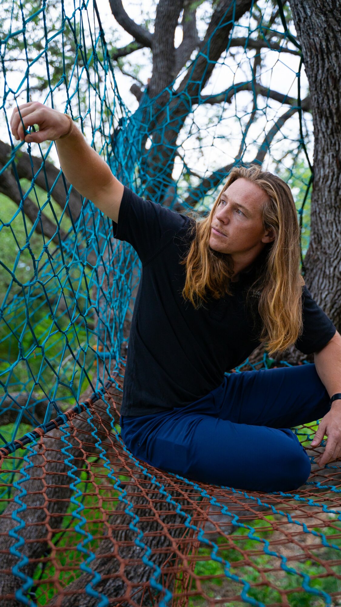 Woman with long hair sitting on a netted structure outdoors, reaching out to touch the net, surrounded by trees.