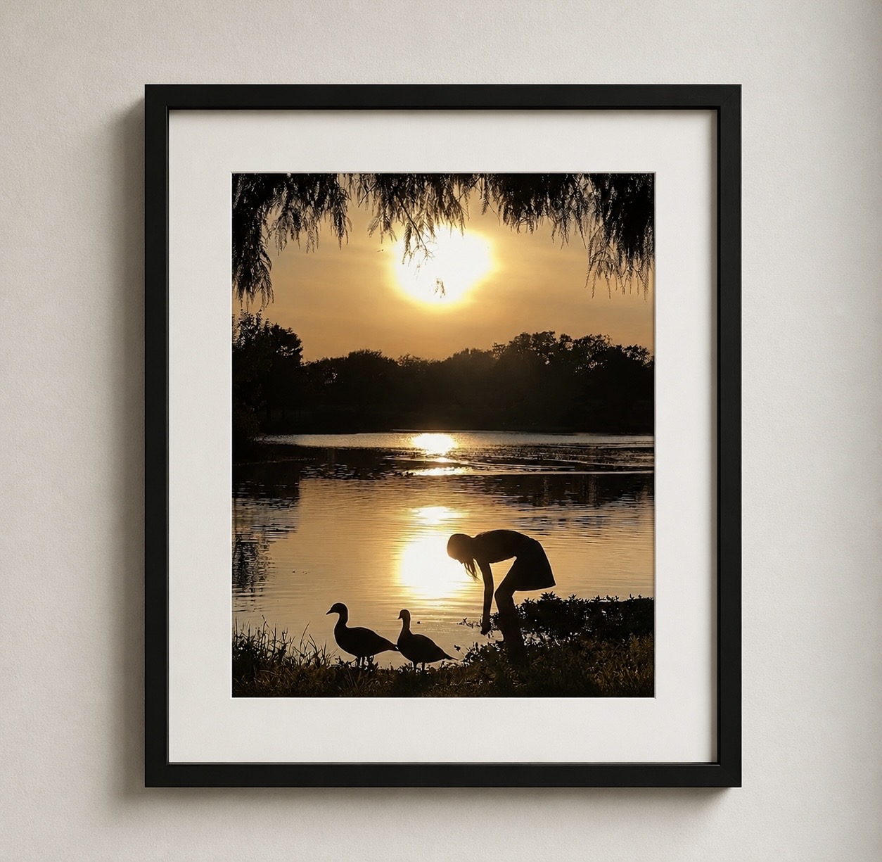 Person bending down near water with two ducks at sunset, trees in background, framed photo, neutral wall