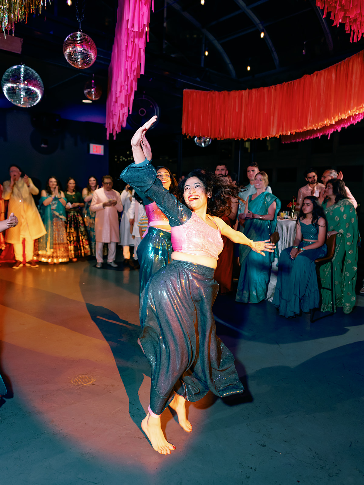 A woman dances in the center of a group, with arms outstretched and one leg raised, in a colorful event space.