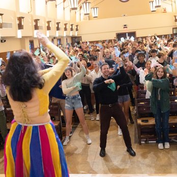 Performer in colorful skirt leading a dance with a large crowd in a hall, people raising hands and smiling.