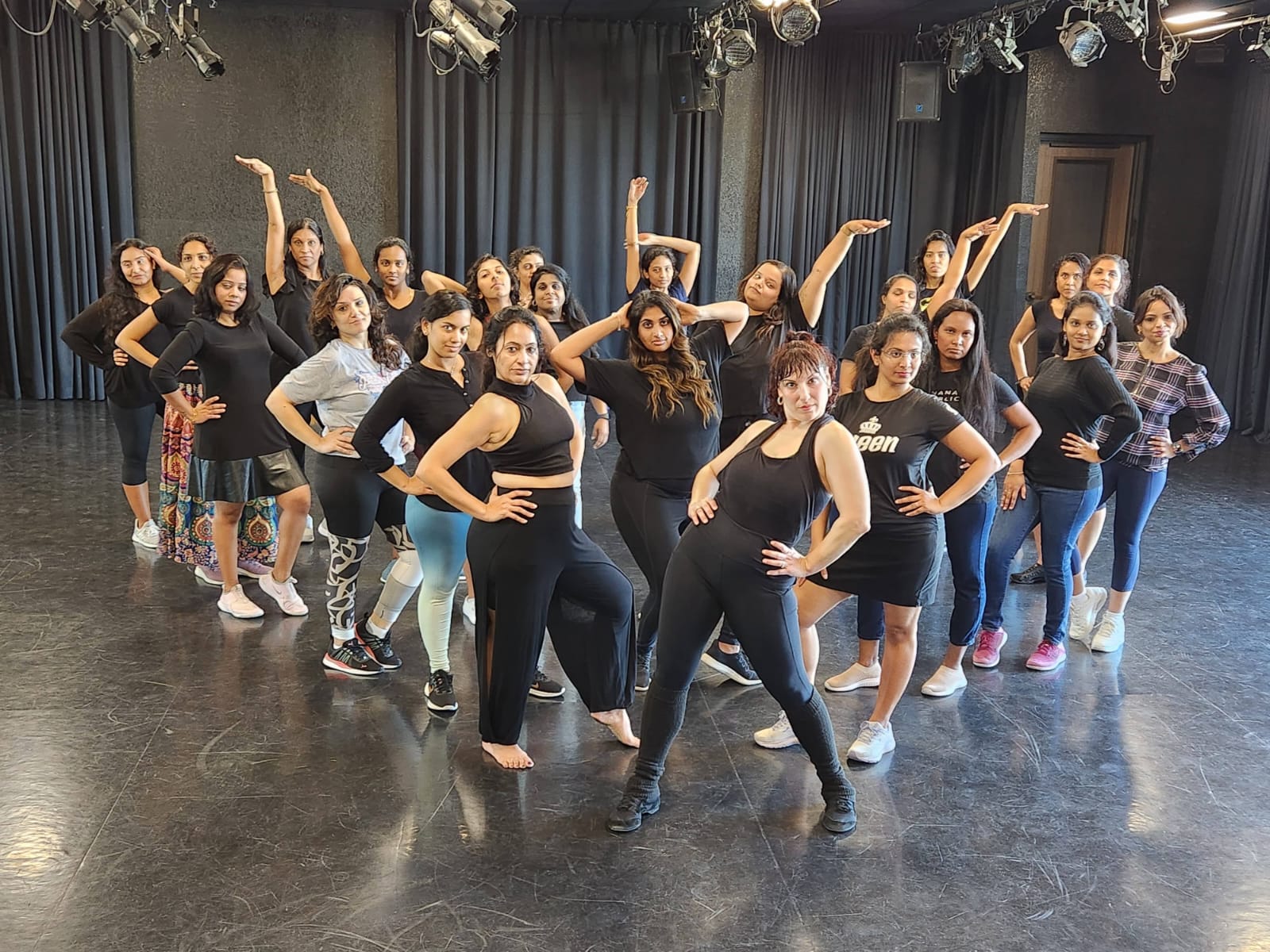 Group of women in black outfits posing on a stage with black curtains, some with hands on hips, others with arms raised.