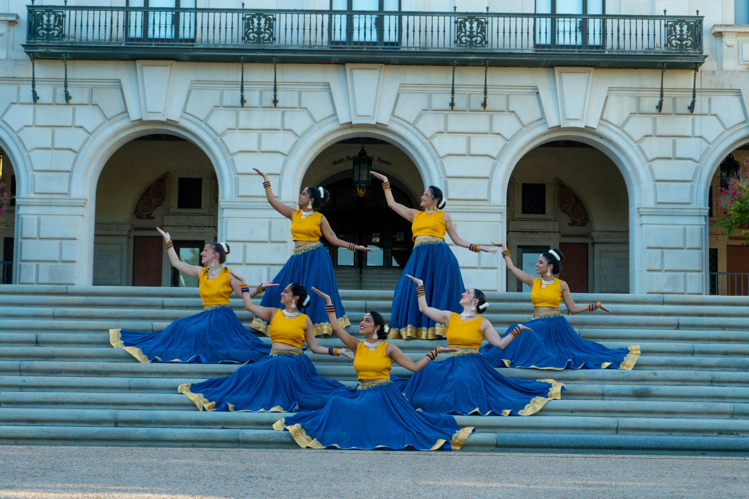 Group of dancers in yellow tops and blue skirts performing on steps in front of a building with arches.