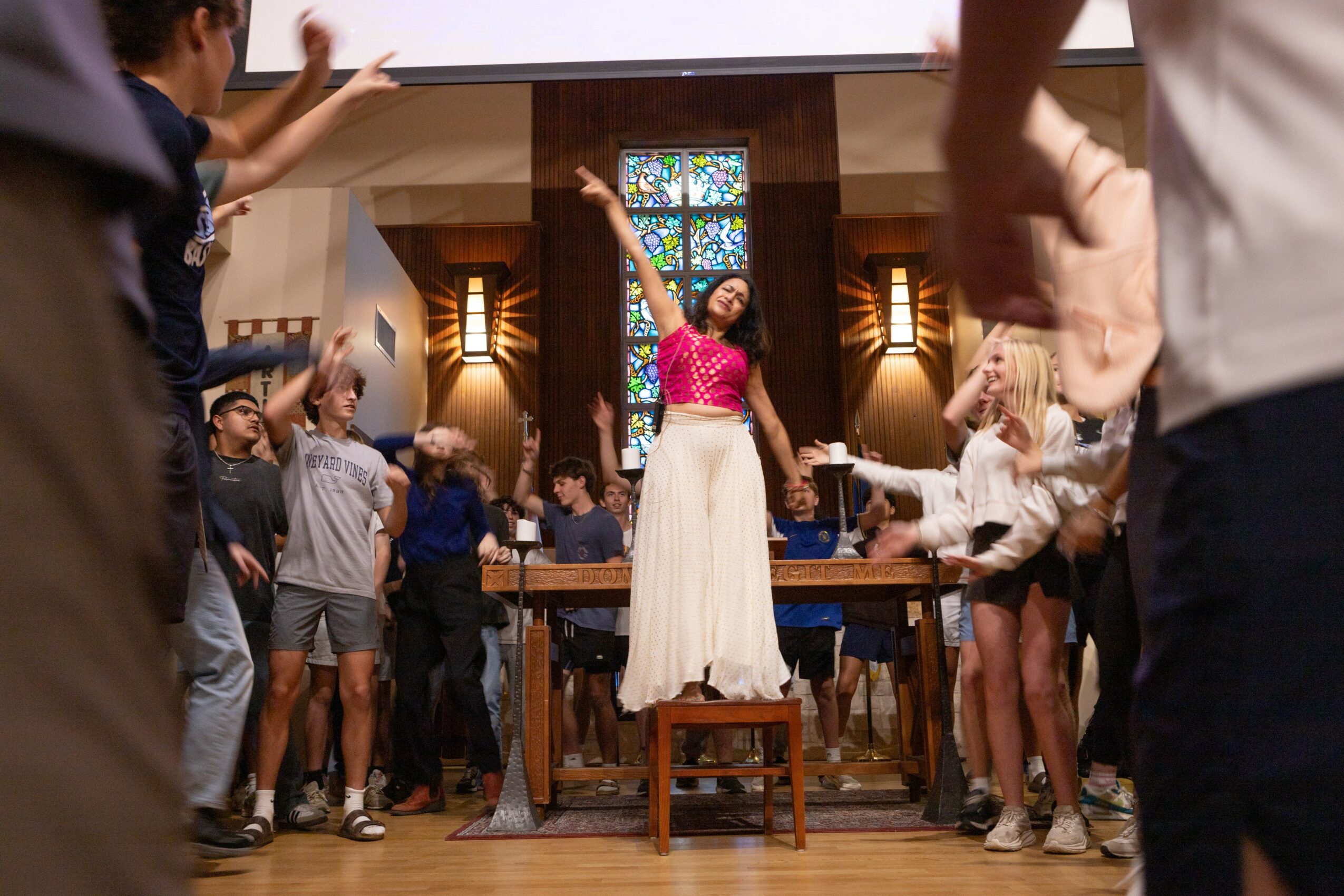 A woman in a pink top and white skirt stands on a chair with arms raised in a church setting, surrounded by people.
