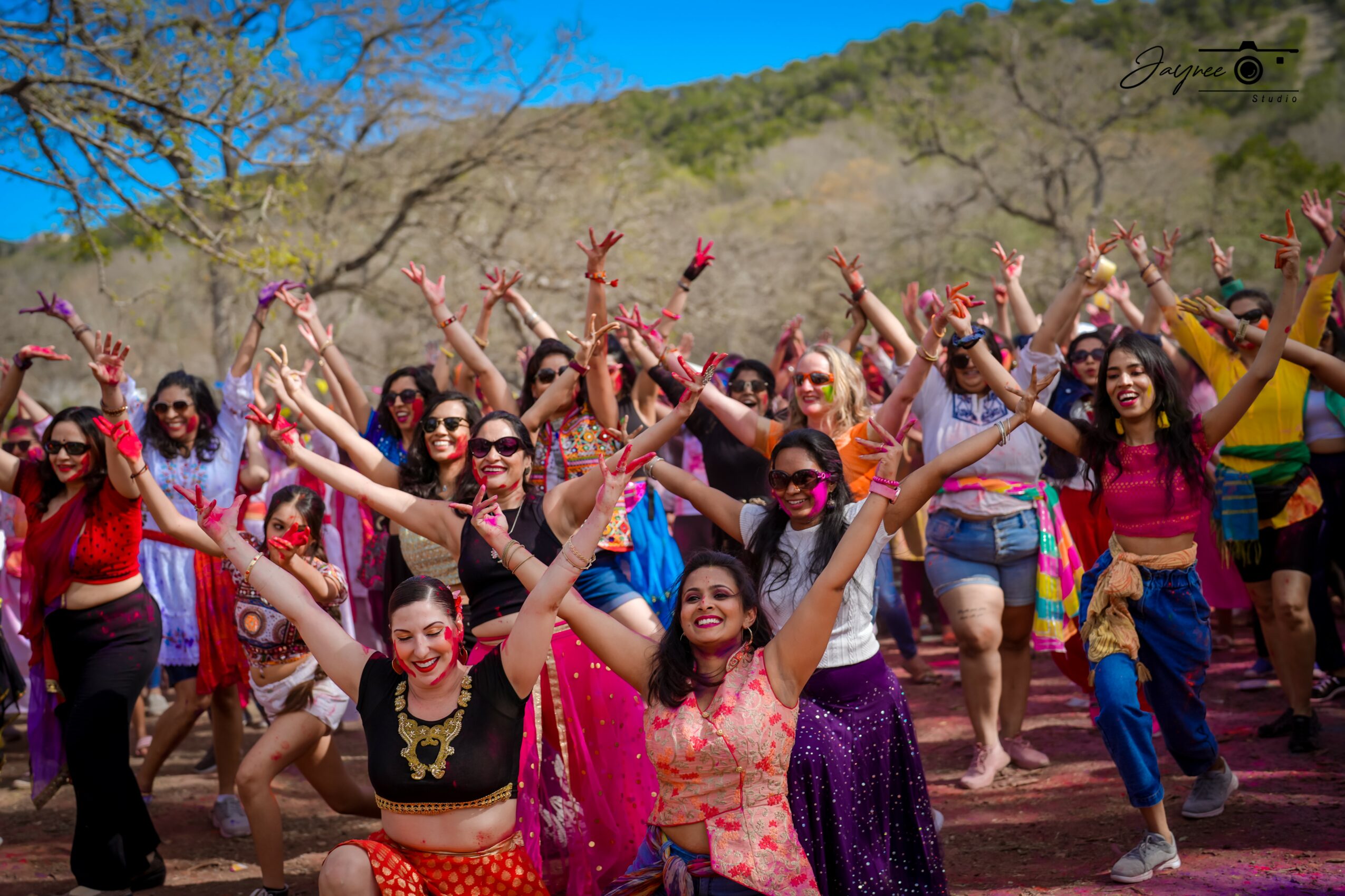Group of women dancing outdoors with arms raised, wearing colorful clothing and sunglasses, in a natural setting with trees and hills.