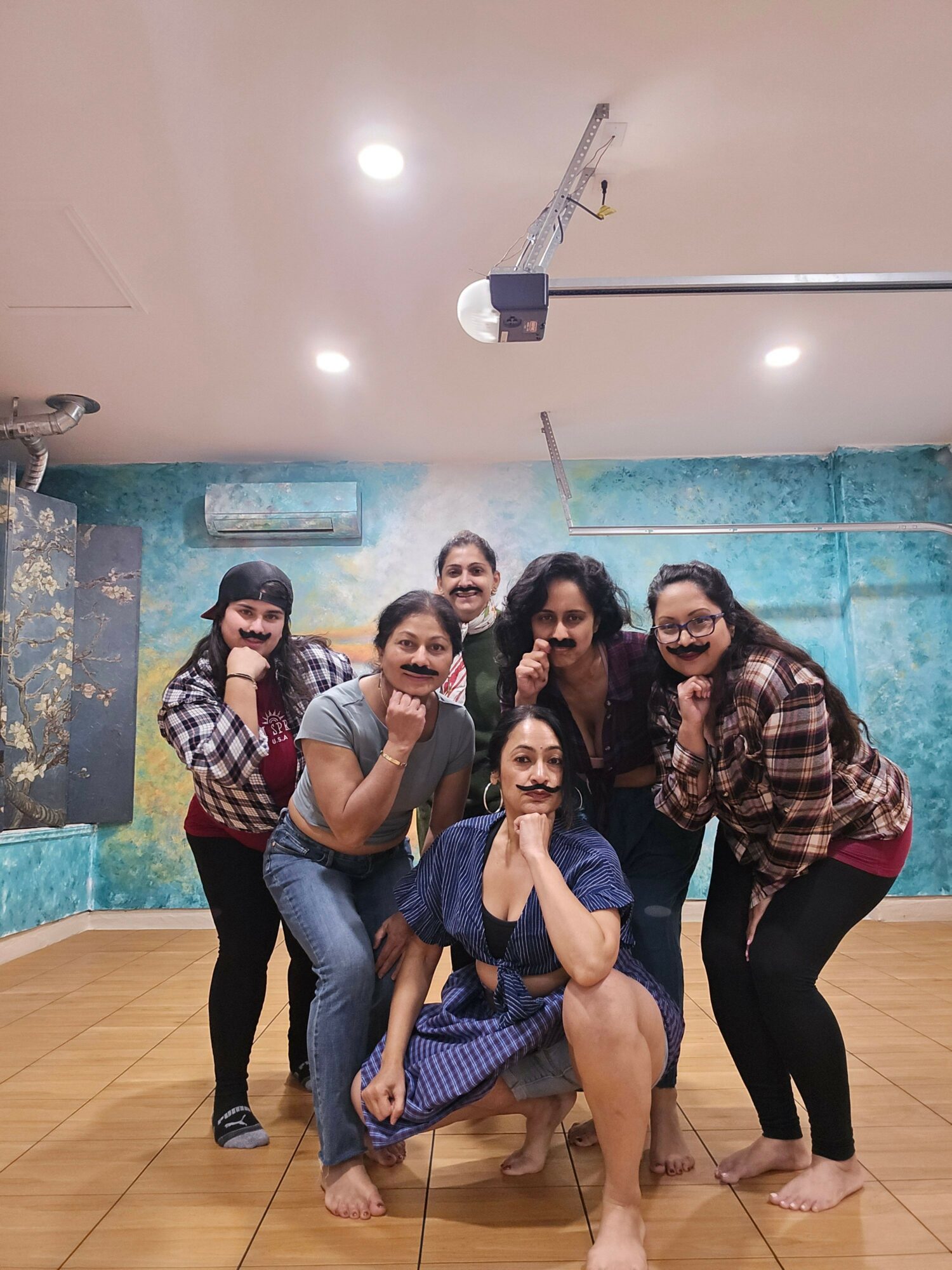 Group of six people posing indoors, some sitting and some standing, with a blue wall and wooden floor.