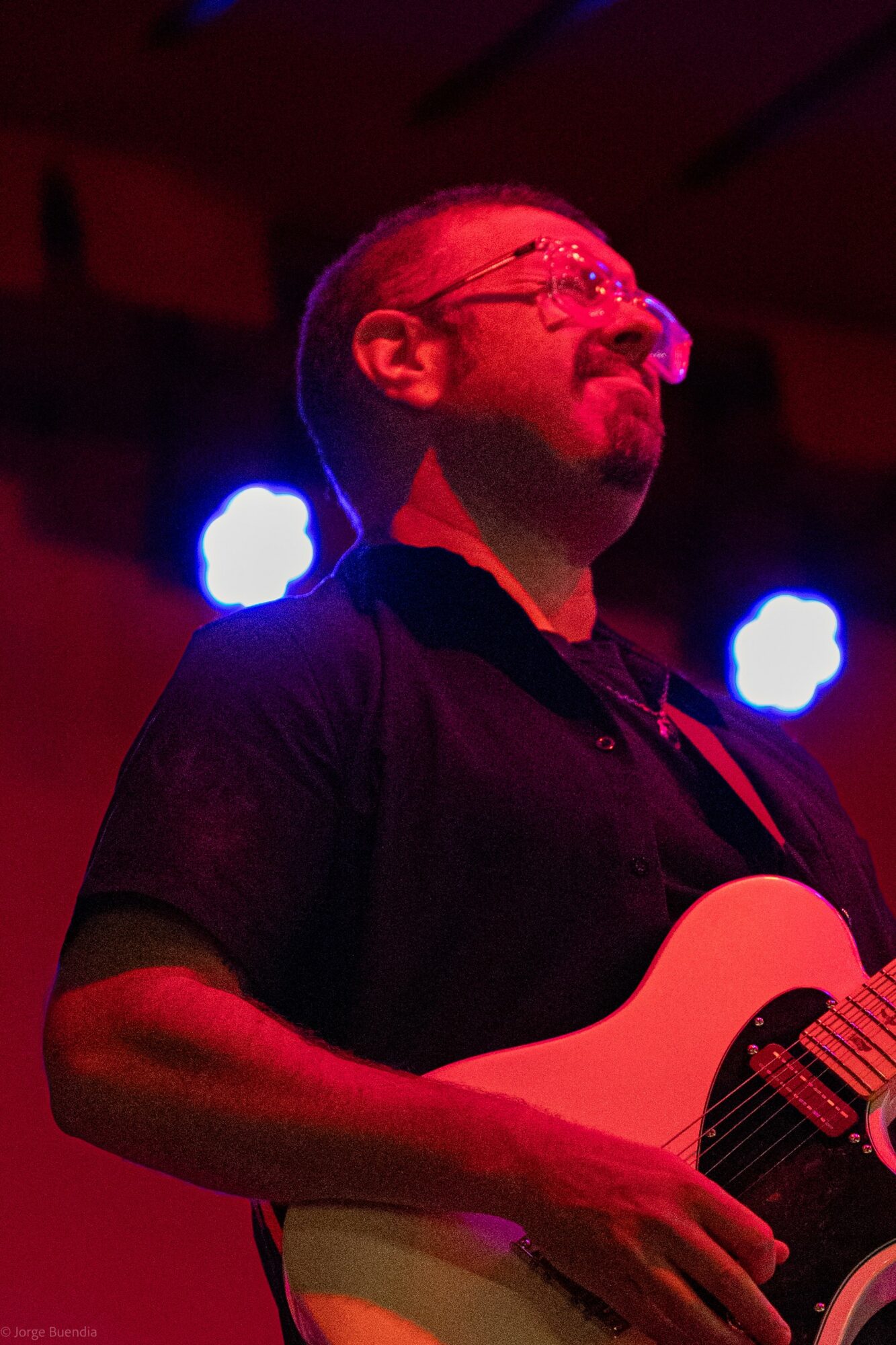 Man playing guitar on stage with colorful lights behind him, wearing glasses and a dark shirt.