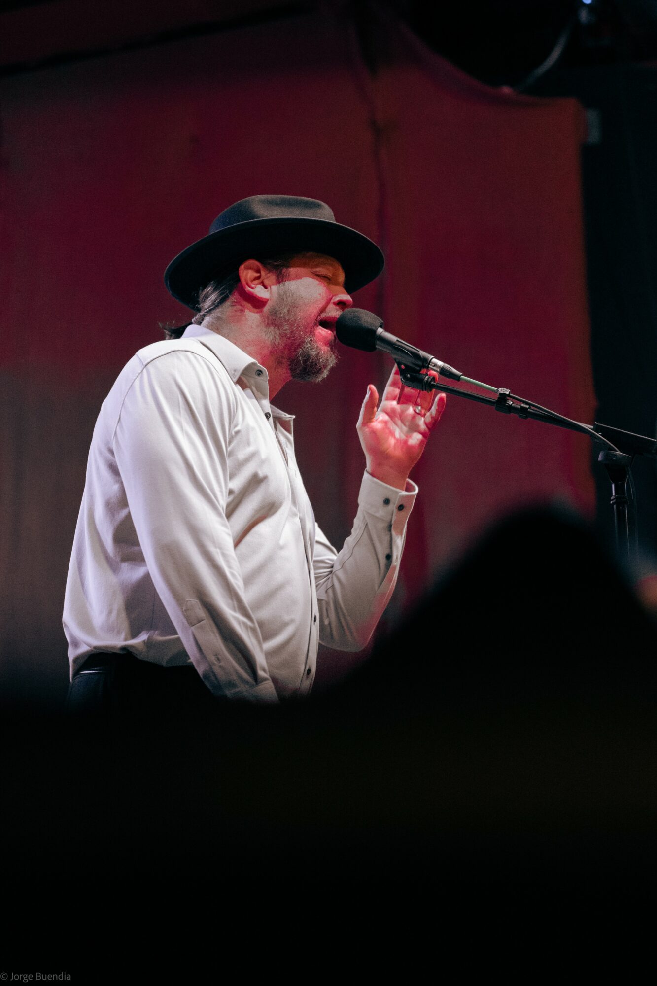 Man singing into microphone, wearing a hat and white shirt, against a dark red background.
