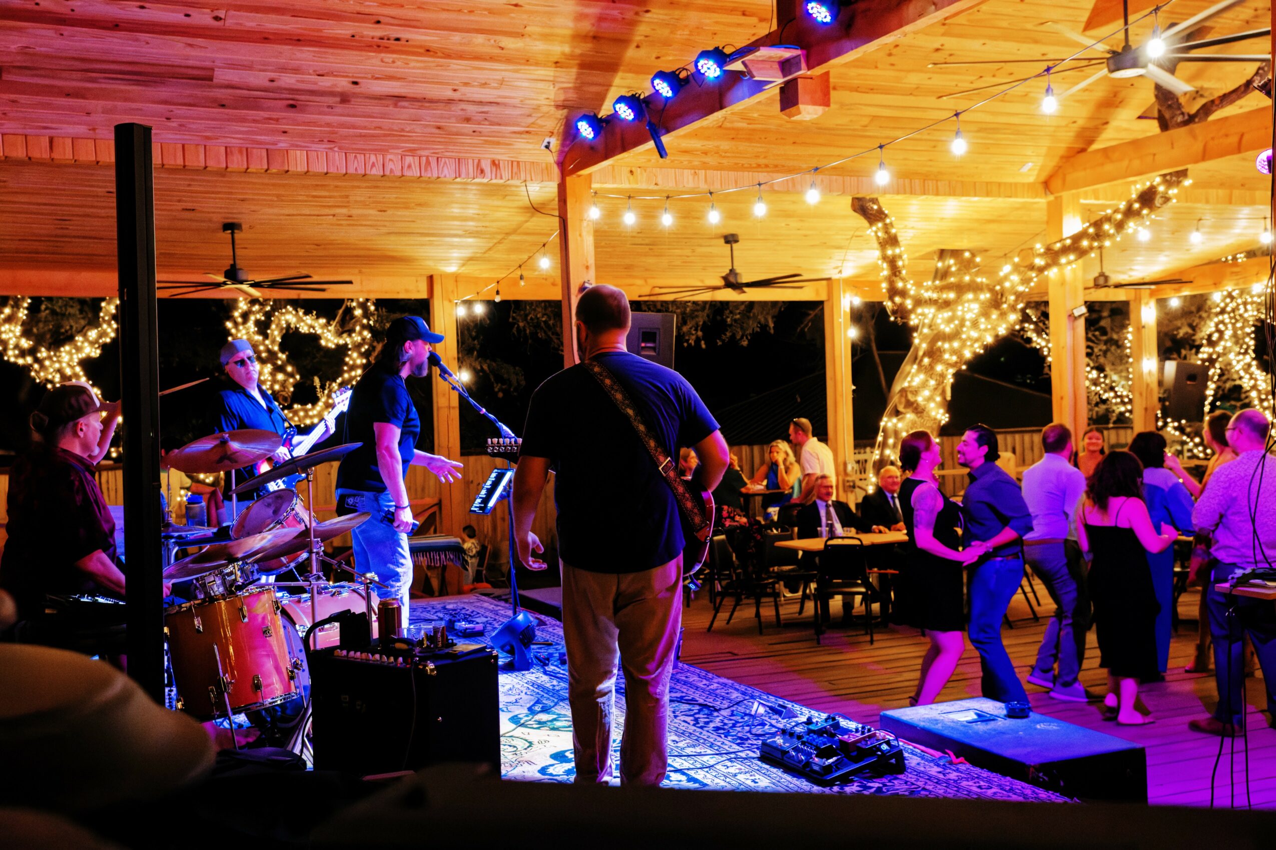 People dancing and a band performing under string lights in a wooden outdoor venue at night.
