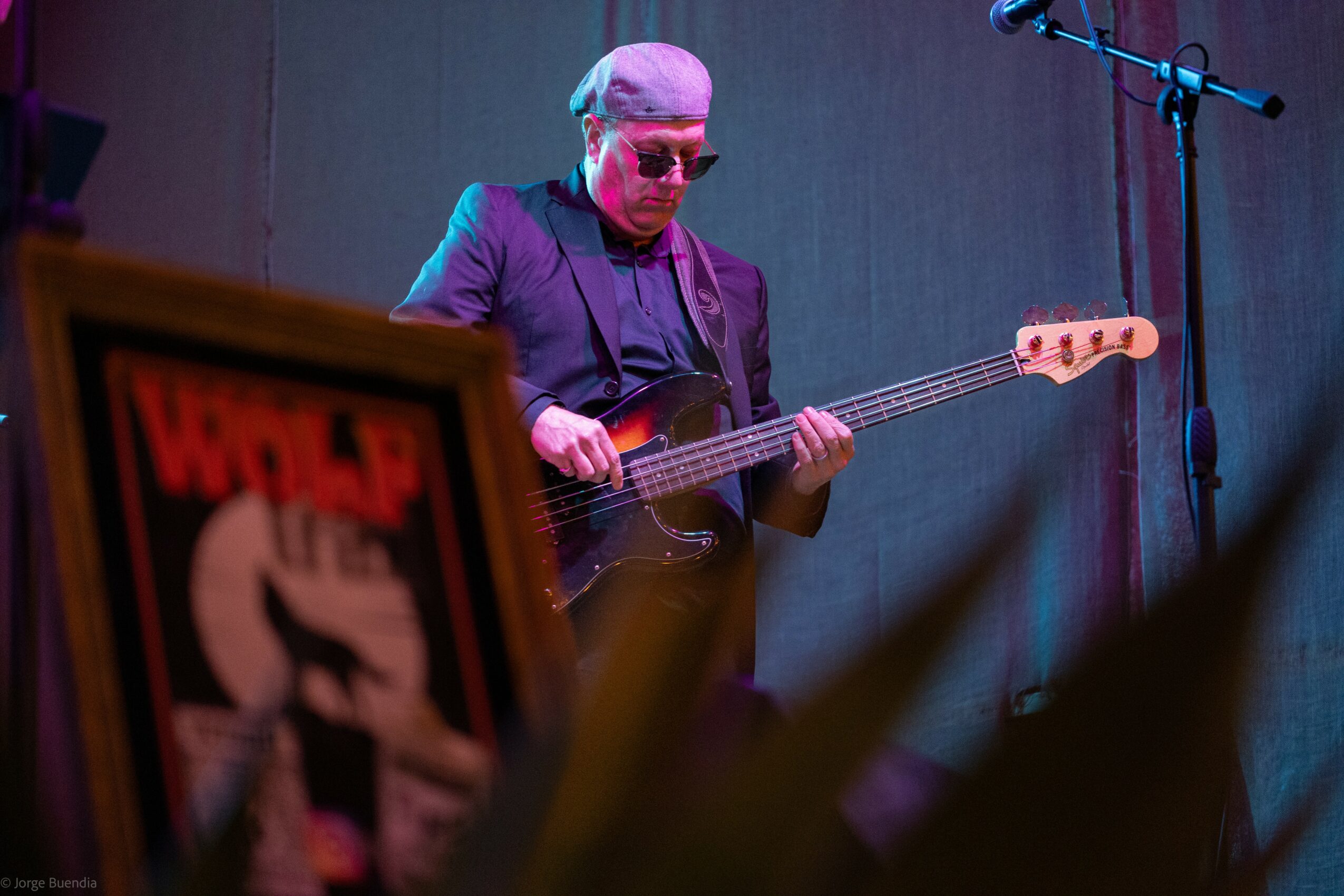 Person playing bass guitar on stage with microphone, background is plain, foreground has blurred objects.