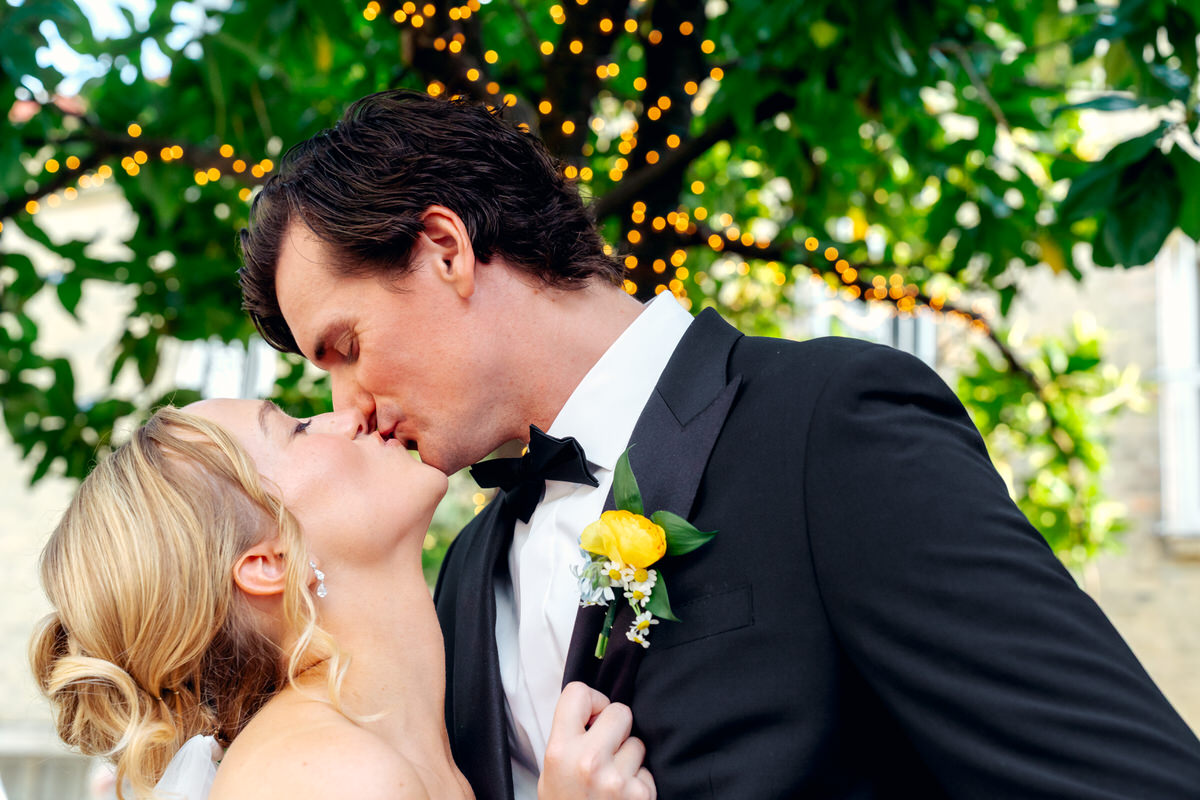 A couple in formal attire sharing a kiss outdoors with greenery and string lights in the background.