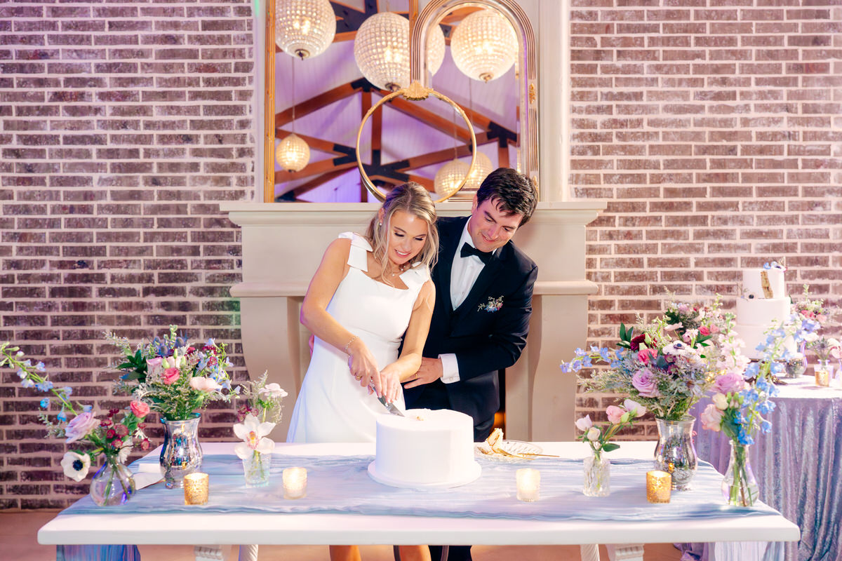 Bride and groom cutting a wedding cake at a decorated table with flowers and candles, brick wall background.