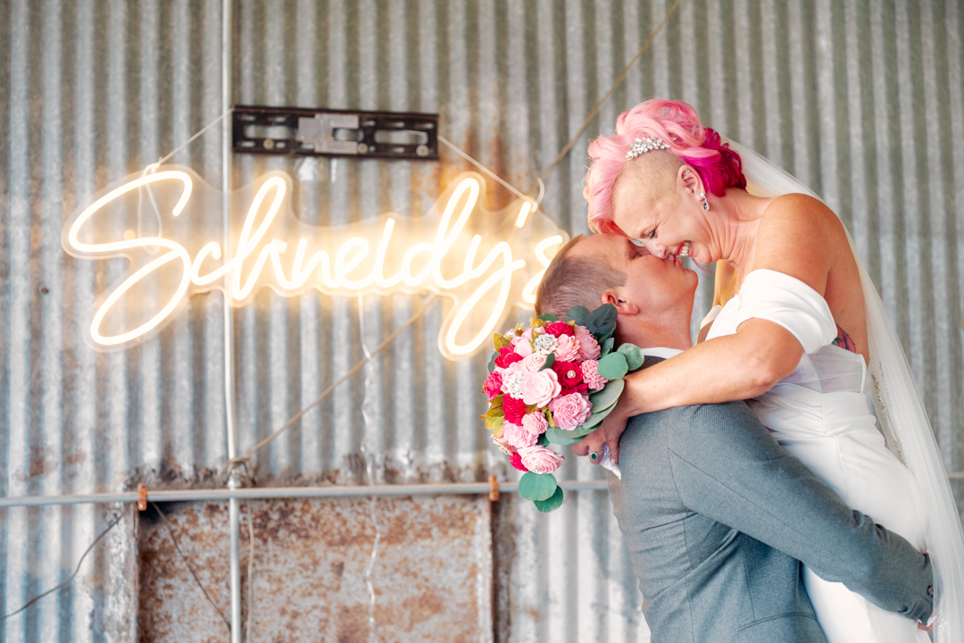 A couple embraces, with the woman holding a bouquet, in front of a neon sign that says 'Scheduled'.