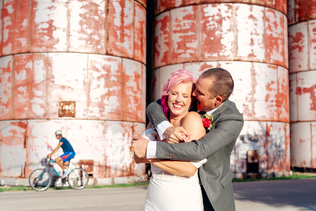 A couple hugging and smiling outdoors with large rusted storage tanks in the background.