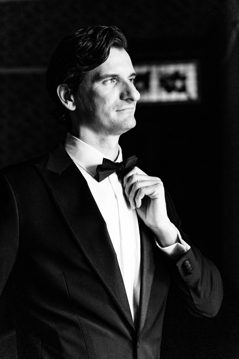 Man in tuxedo adjusting bow tie, looking to the side, in black and white.