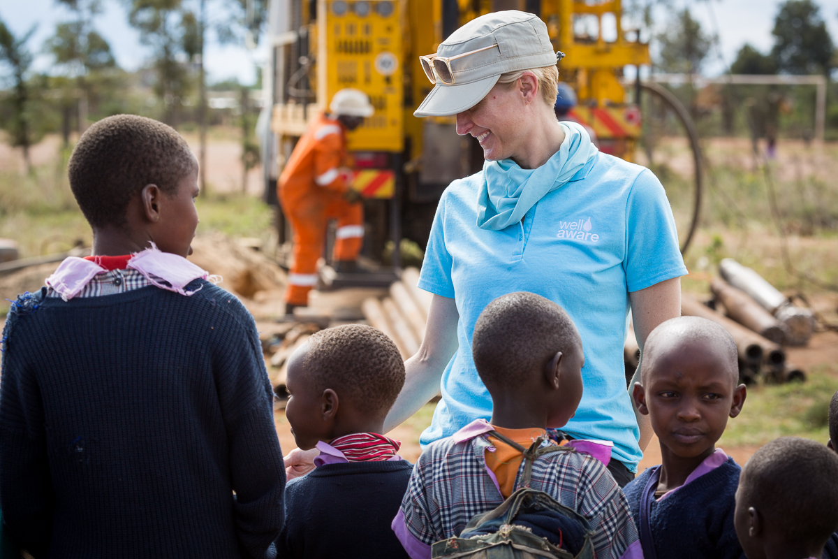 A woman in a light blue shirt and cap talking to children outdoors with construction equipment in background.