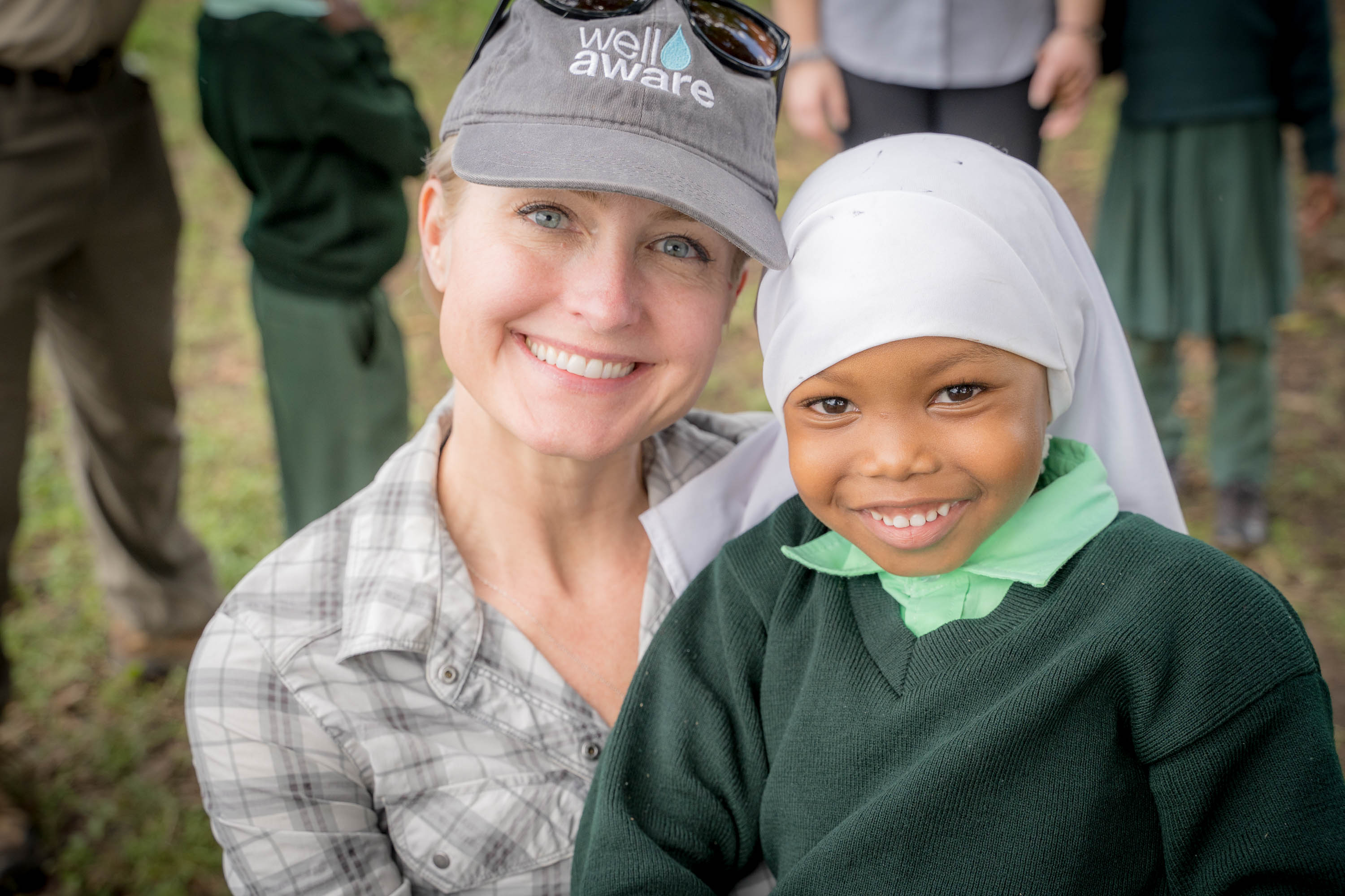 Smiling woman and girl in head coverings outdoors, group in background, natural setting, daytime.