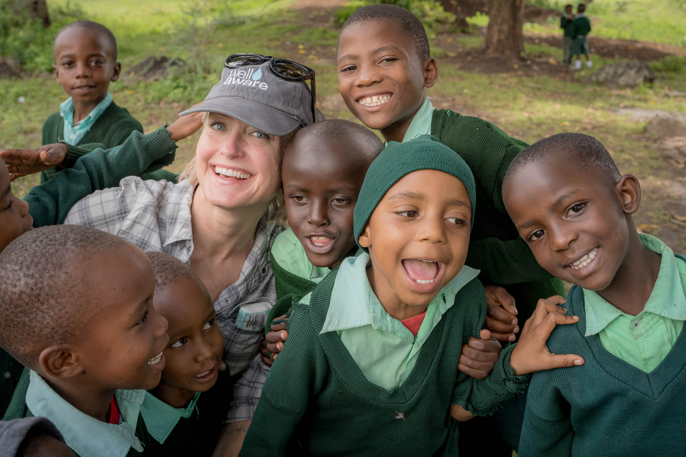 Group of children and a woman smiling outdoors, some children wearing green uniforms and a woman with a cap.