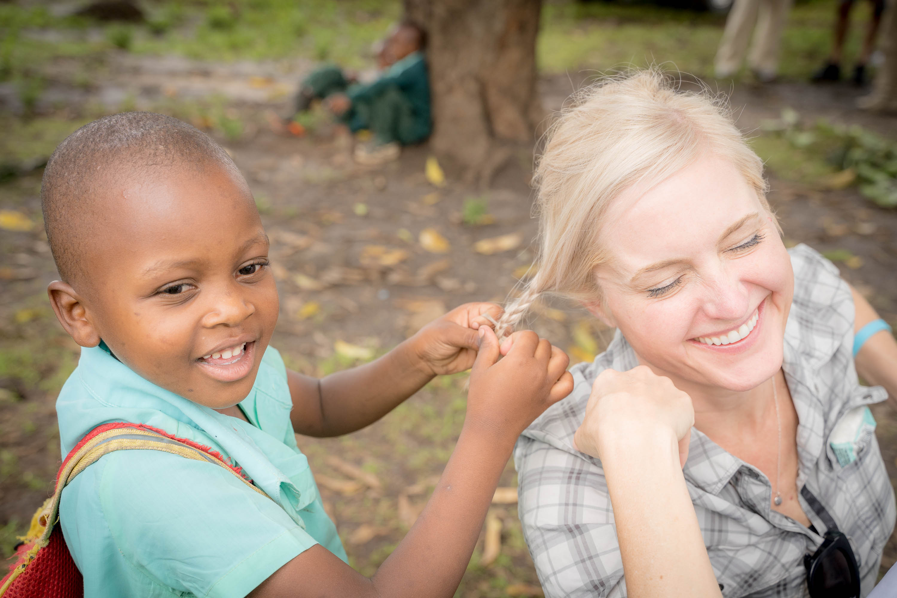 A young boy with a backpack smiling and pulling blonde woman's hair outdoors, trees and ground visible.